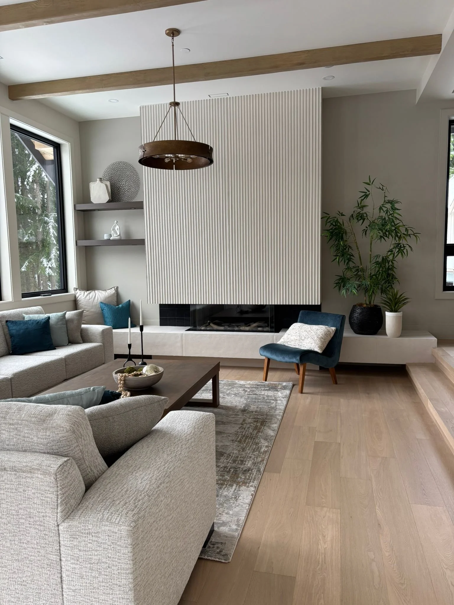 Modern living room with beige sofa, blue and teal pillows, wooden coffee table, fireplace, large potted plant, window, and ceiling light fixture.