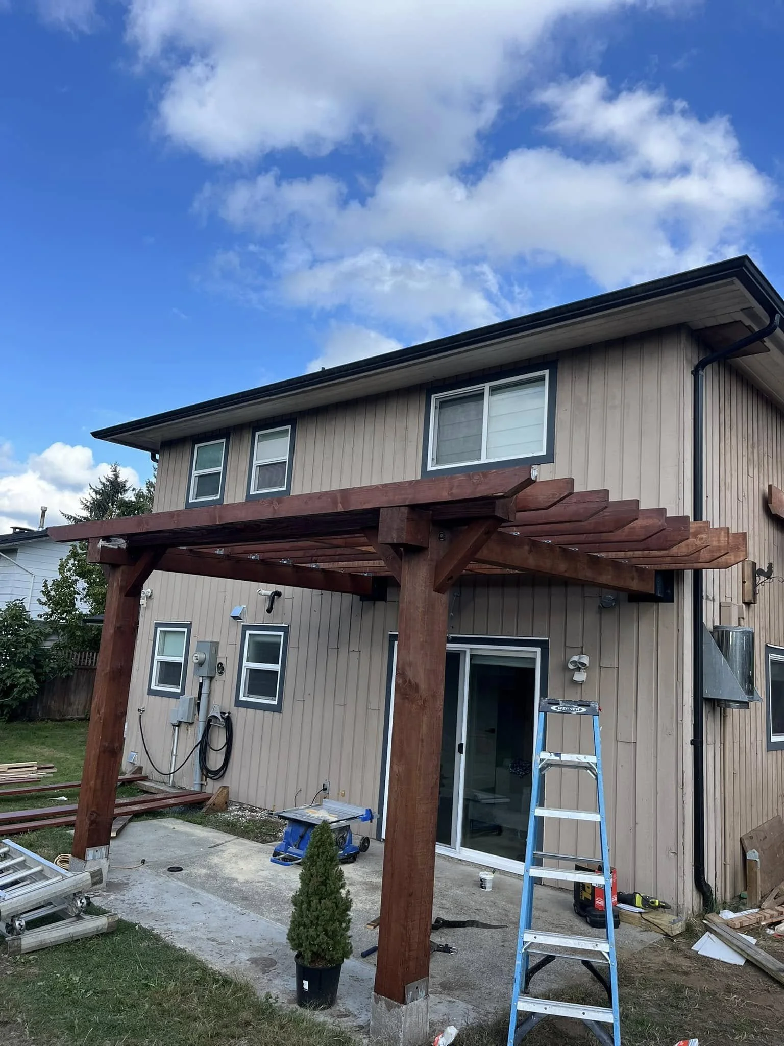 Backyard of a two-story house with a partially built wooden patio or pergola structure, a ladder, tools, and construction materials visible.