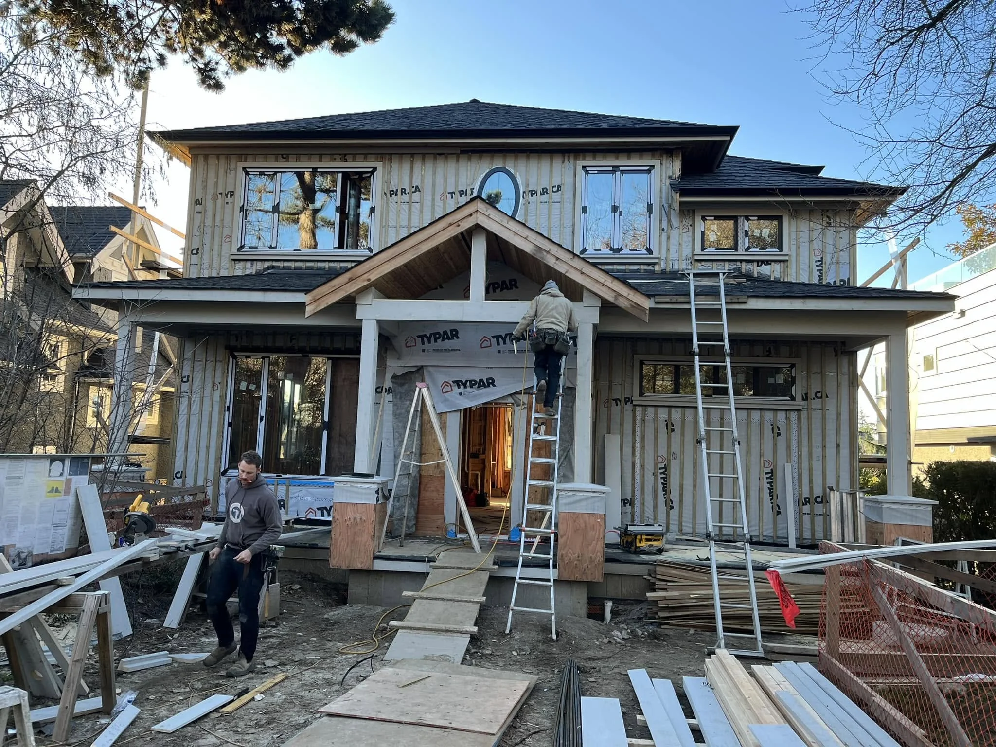 Construction workers building a two-story house, with ladders, construction materials, and tools around the unfinished exterior, and the house being covered in weatherproofing material.