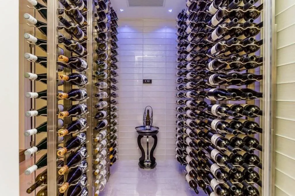 A wine cellar with two walls of horizontal wine racks filled with bottles, a small table in the middle, and a white tiled wall at the back.