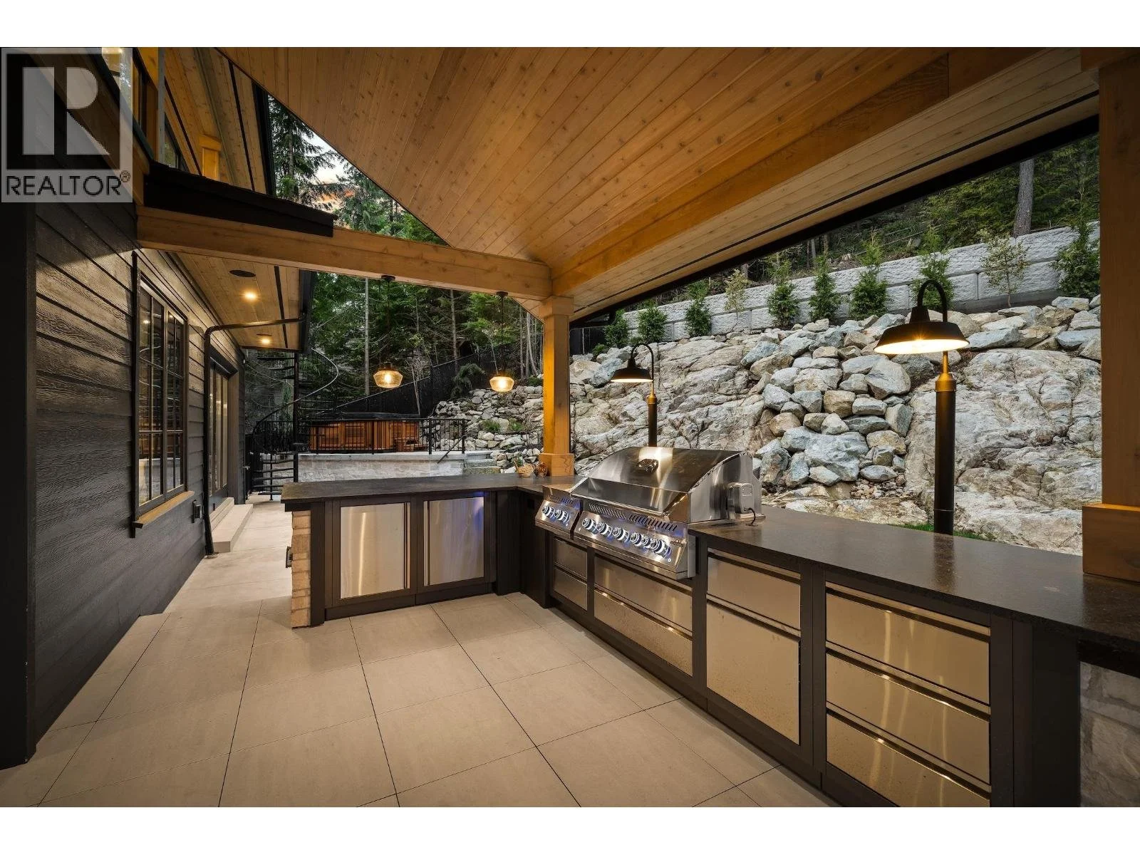 Outdoor kitchen with stainless steel appliances, dark cabinets, and a tiled floor. It has a wooden ceiling and is situated against a rocky hillside with small trees, illuminated by black outdoor lamps.
