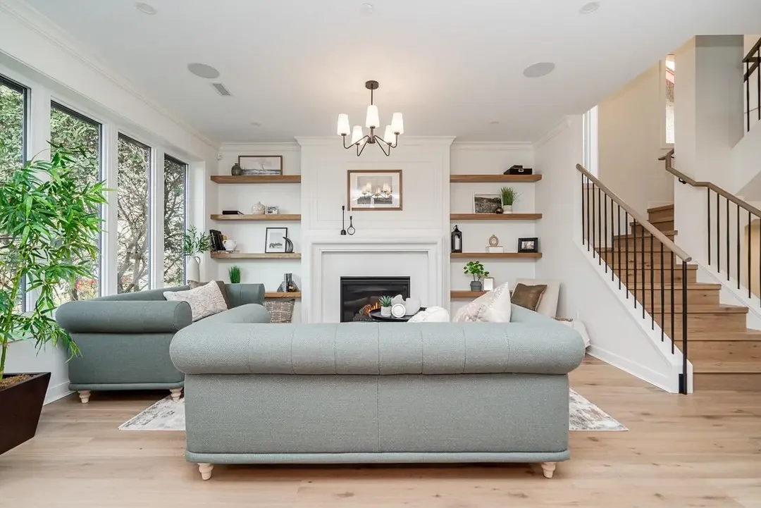 Bright living room with large windows, two light green sofas, a fireplace, floating shelves with decorations, a staircase with wooden steps and black railing, and a chandelier, with hardwood flooring and indoor plants.