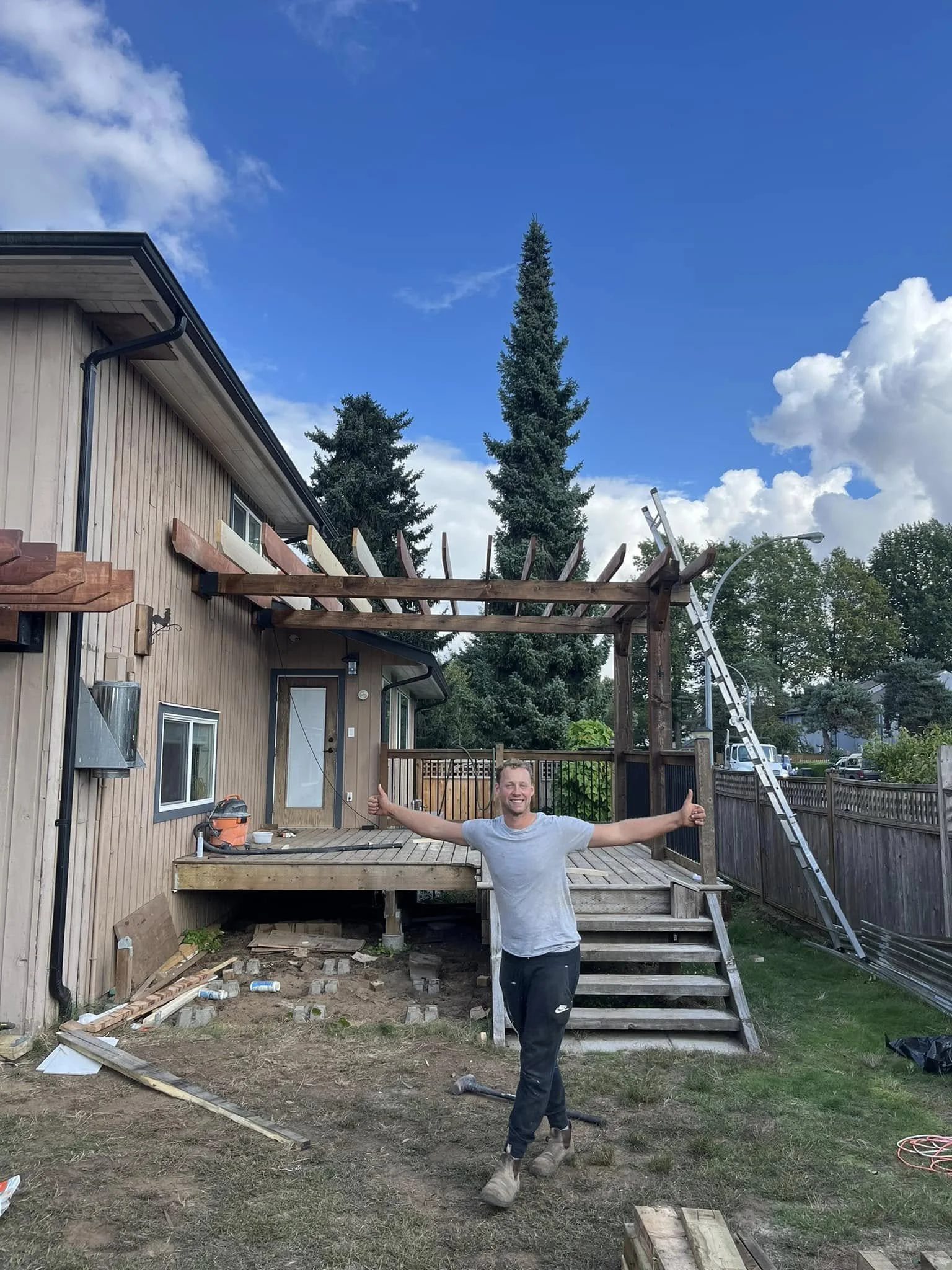A man standing in a backyard giving a thumbs up with a completed wooden deck under construction and a person ladder in the background.