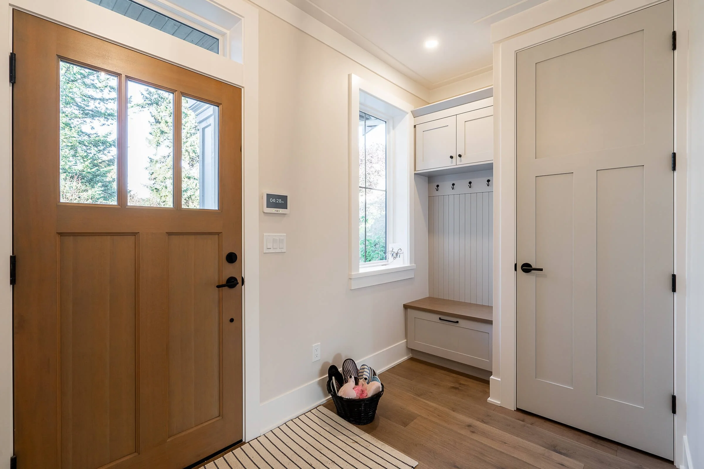 Entrance hallway with a wooden front door featuring windows, a window with a windowsill, a bench with a drawer, hooks for coats, a basket with shoes, a striped rug, and a digital clock on the wall.
