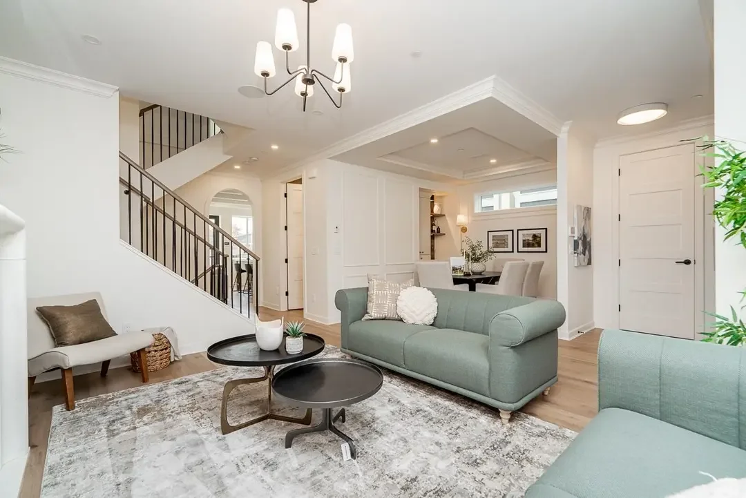 Bright living room with white walls and light wood flooring, featuring a white sofa, an upholstered green sofa, a small armchair with a dark pillow, a black coffee table with decorative items, a white area rug, a staircase with black railings, and a 