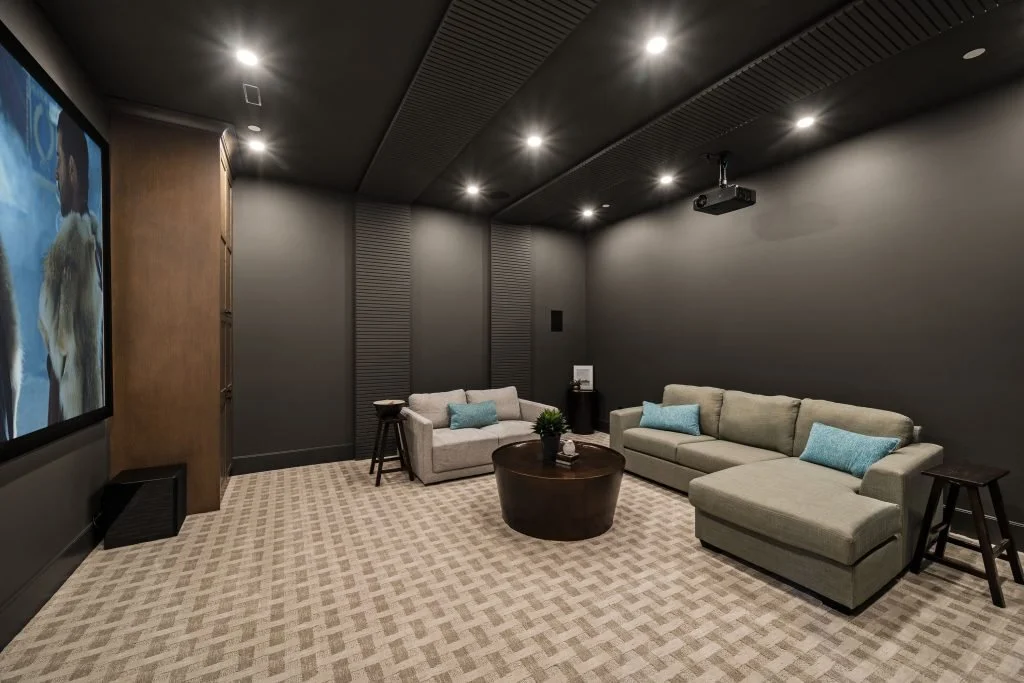 A home theater room with black walls and ceiling, a large screen on the left displaying a movie, beige sofas with blue pillows, a round coffee table, and small side tables, with a projector mounted on the ceiling.