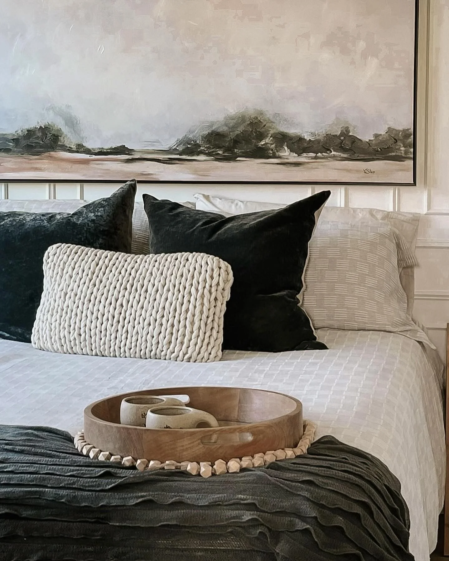 A neatly made bed with a white textured bedspread, multiple decorative pillows in black, white, and cream, a round wooden tray with three small ceramic cups, and abstract wall art above the headboard.