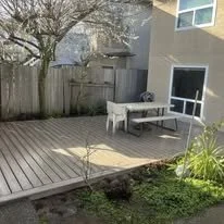 Backyard patio with wooden deck, a white bench, a small table, a white dog, and surrounding plants and trees.