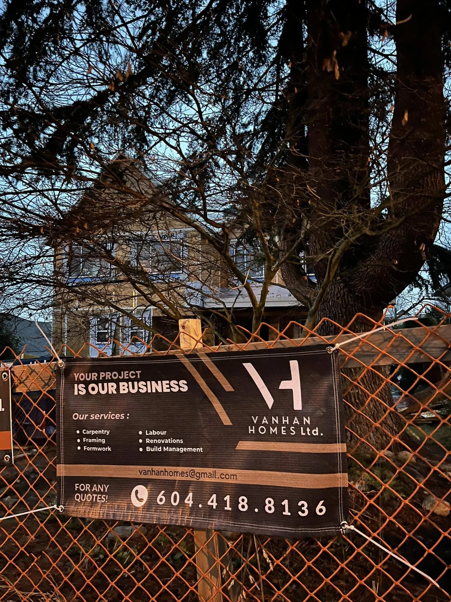 A construction site sign attached to a chain-link fence in front of a house with large trees. The sign advertises Vanhan Homes Ltd., offering services like carpentry, framing, formwork, labor, renovations, and build management. The contact email is v