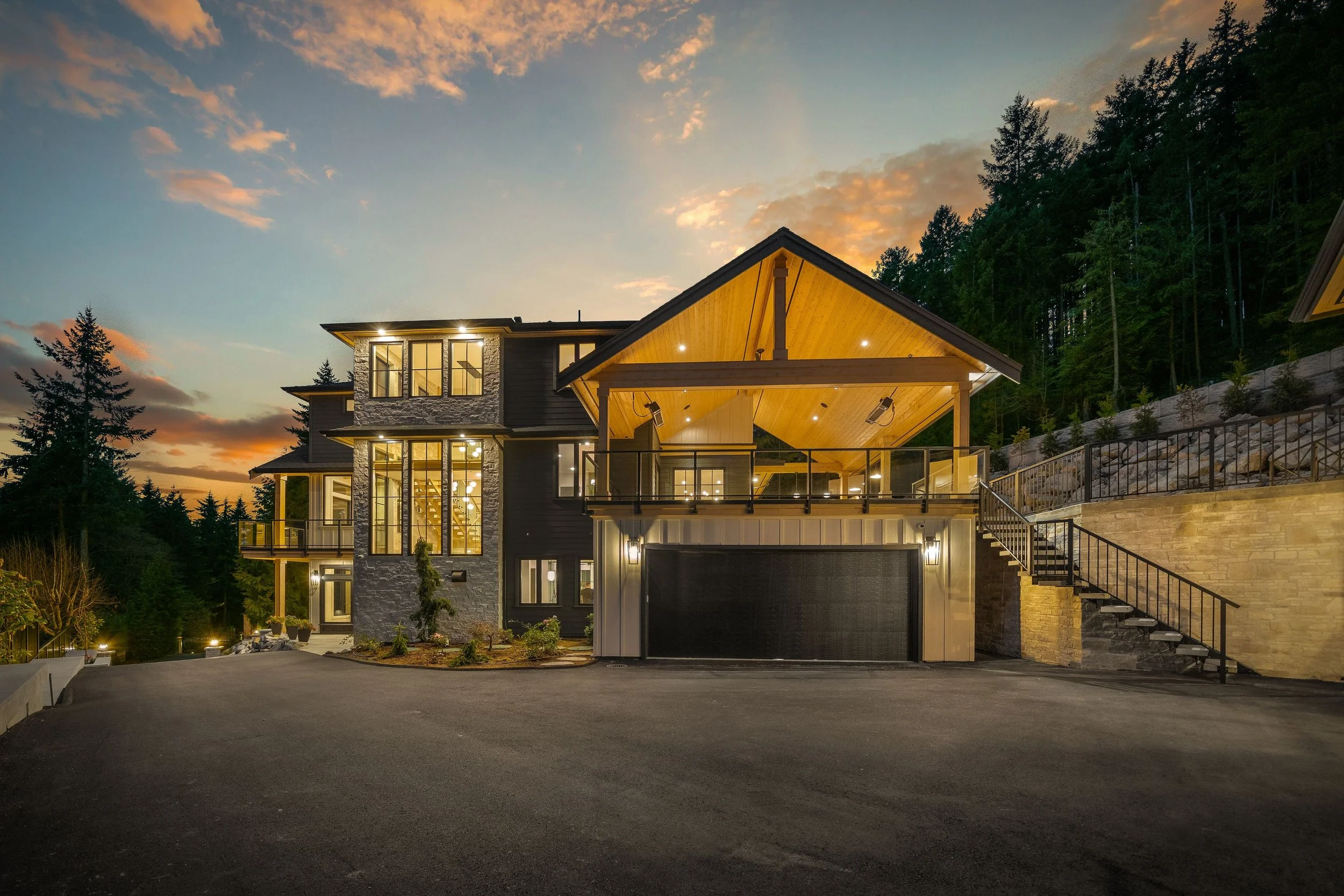 Modern multi-story house at dusk with large windows, an attached garage, and a wooden covered balcony, surrounded by trees.