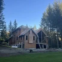 A two-story wooden house under construction surrounded by trees and clear blue sky.