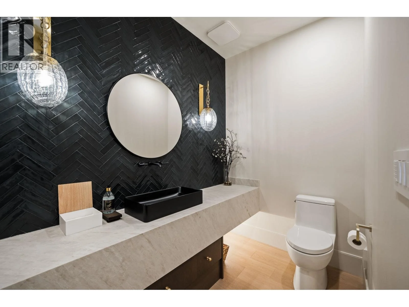 A modern bathroom with black herringbone tile wall, round mirror, black vessel sink, wooden and white storage tray, soap dispenser, white toilet, and decorative branch arrangement on a beige marble countertop.