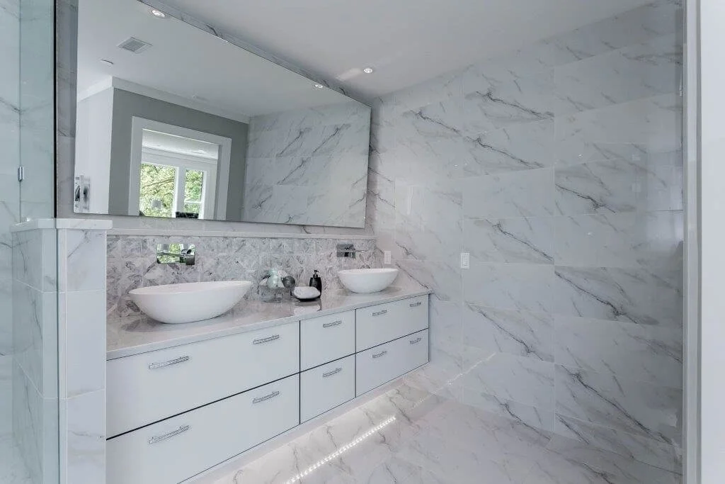 Modern bathroom with white marble walls and floors, a large mirror, a double vanity with two white vessel sinks, and a window showing greenery outside.