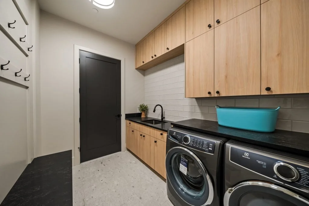 Laundry room with wooden cabinets, a black countertop, a sink with a black faucet, a potted plant, a blue basin, and two front-loading washing machines.