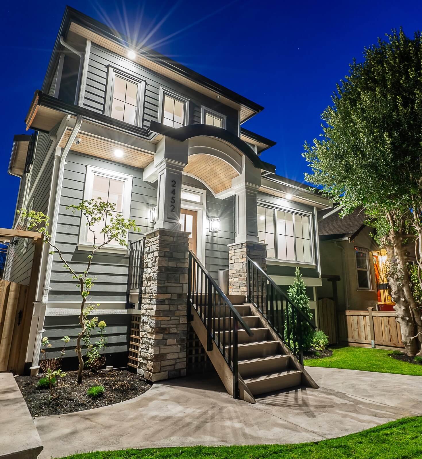 Night view of a modern two-story house with exterior lighting, steps leading to the front door, and landscaped yard with trees and grass.