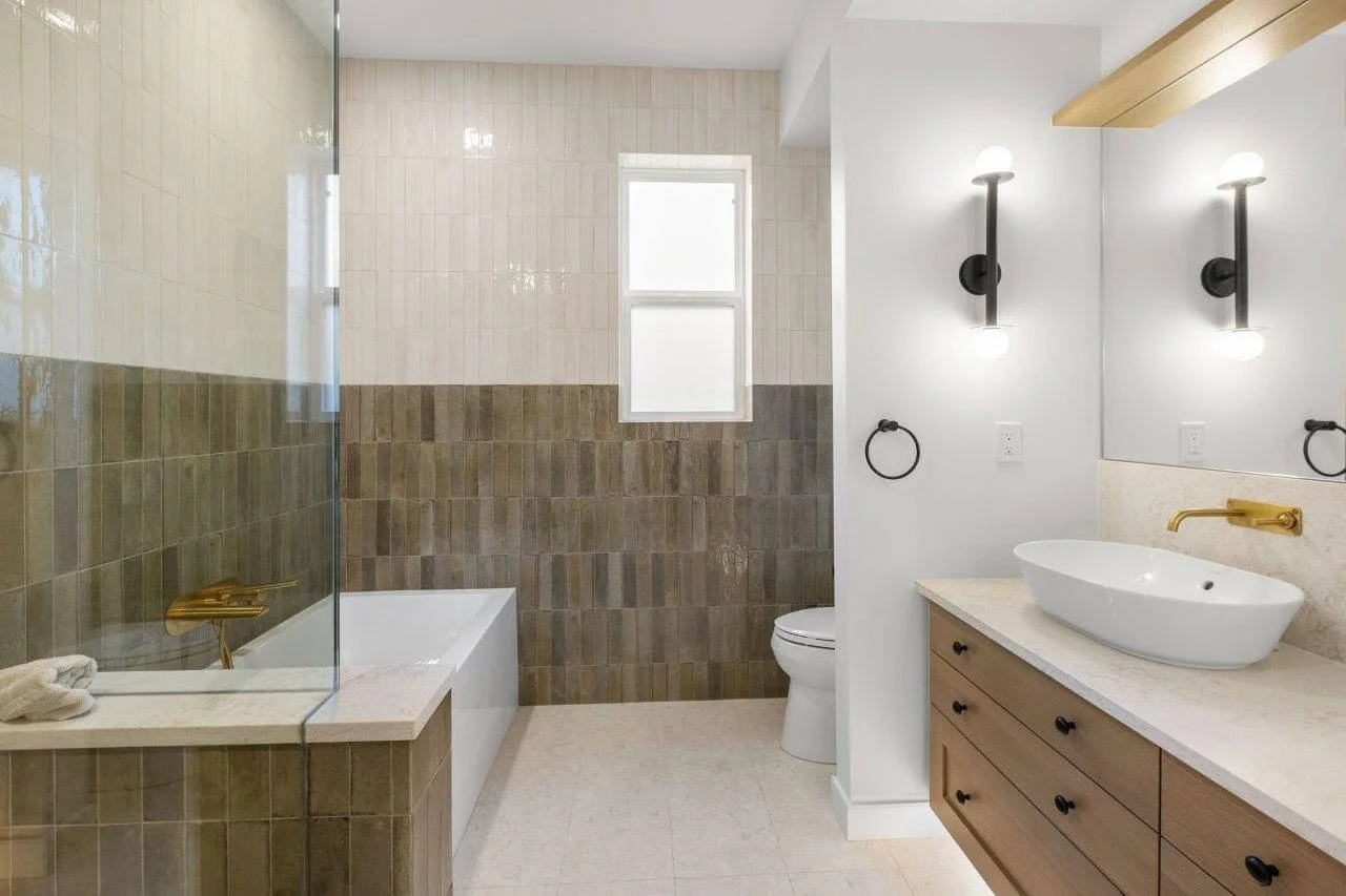 Modern bathroom with a glass-enclosed shower, bathtub, toilet, and a vanity with a vessel sink and black fixtures, featuring a mix of neutral tiles and wooden accents.