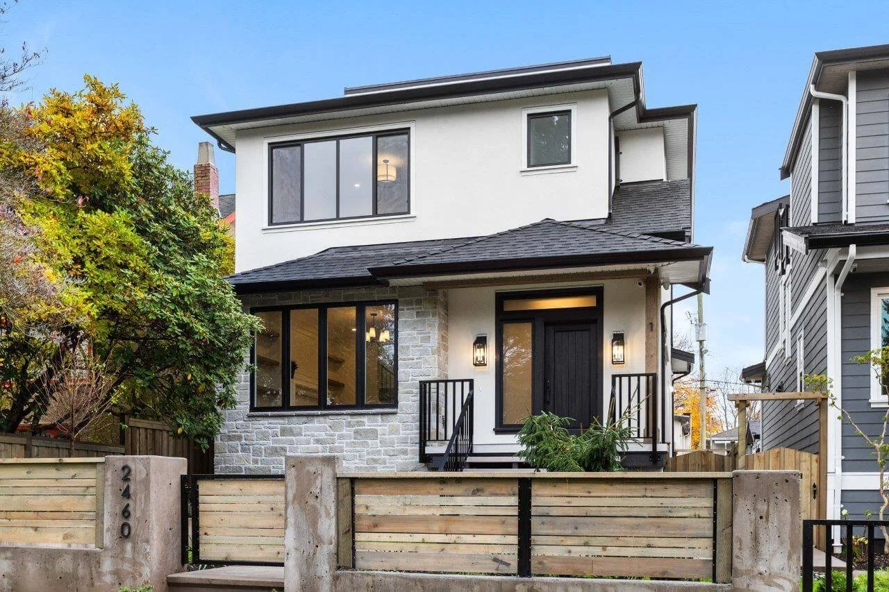Modern two-story house with a black front door, large windows, stone accents, and a small front porch, surrounded by a wooden fence and greenery.