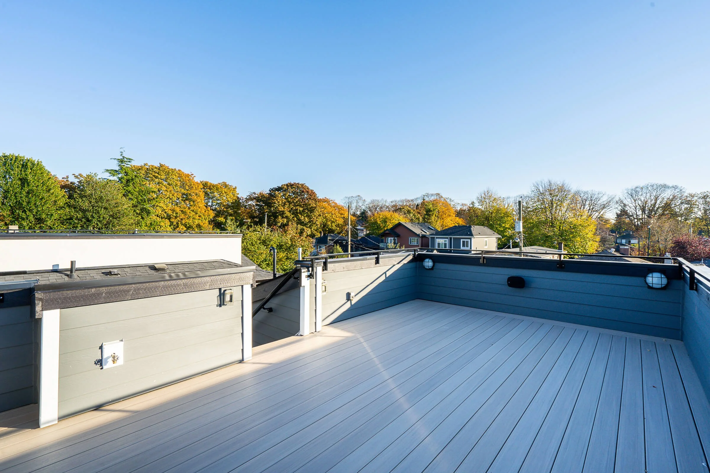 View of a rooftop terrace with a light gray wooden deck, bordered by gray painted walls and black handrails, overlooking a neighborhood with trees and houses under a clear blue sky.