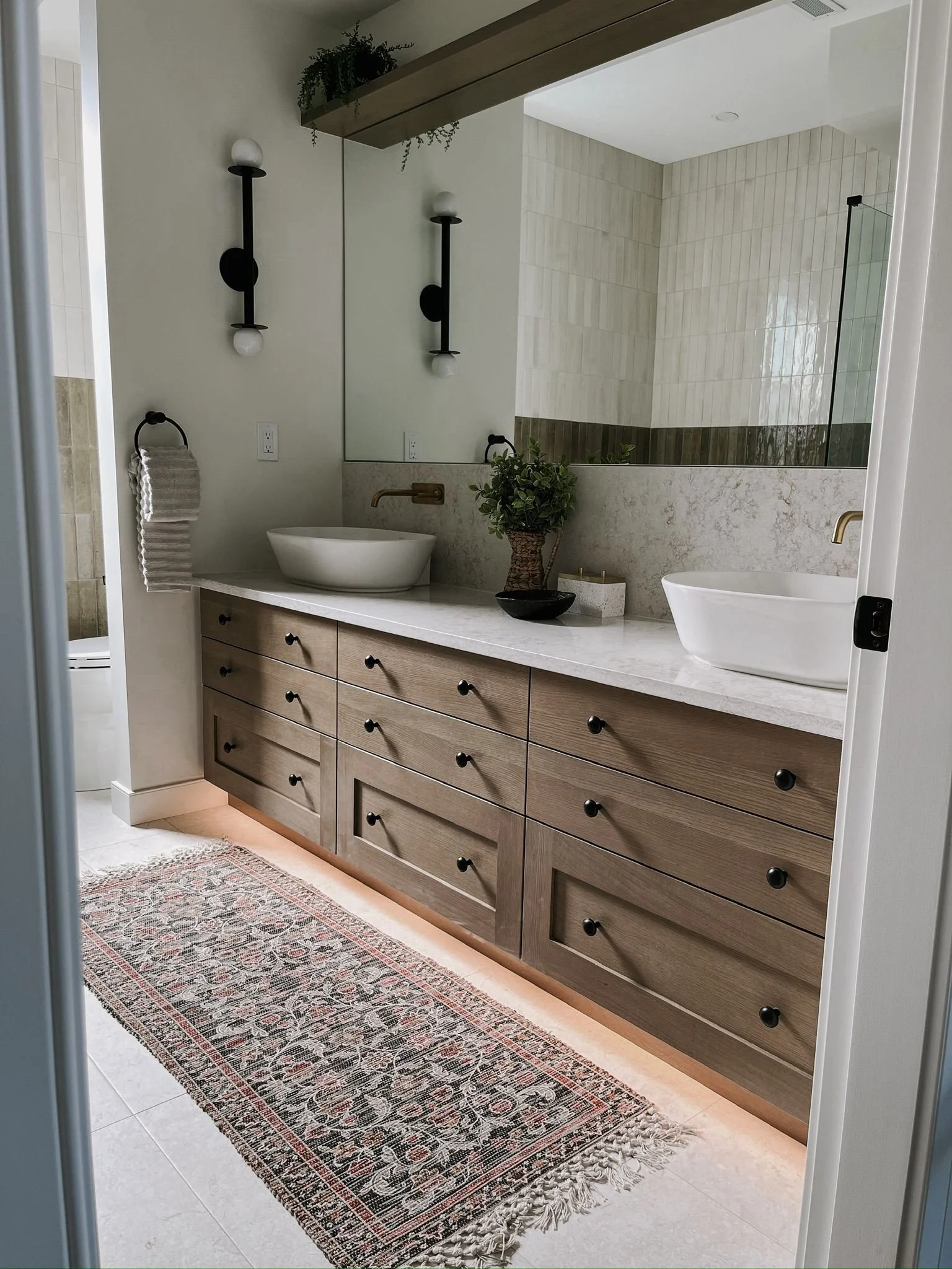 Modern bathroom with a wooden vanity with nine drawers, two white vessel sinks, black hardware, large mirror, wall-mounted lights, a potted plant, a rug, and a shower with glass enclosure.