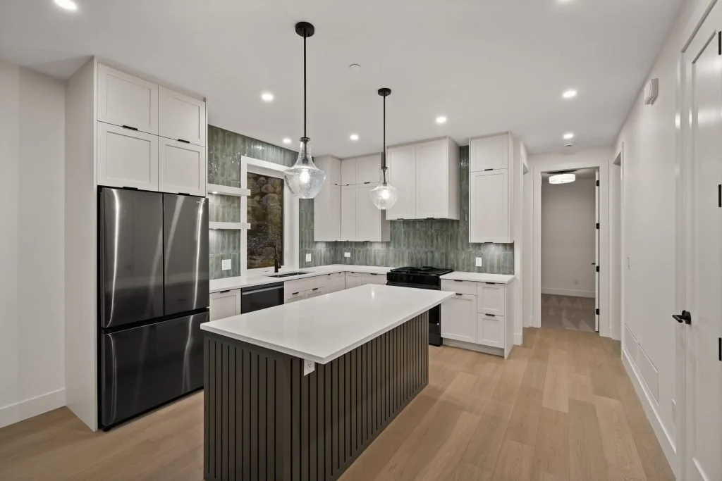 Modern kitchen with white cabinets, stainless steel refrigerator, island with white countertop and dark panel, green tiled backsplash, black stove, pendant lights, and hardwood floors.