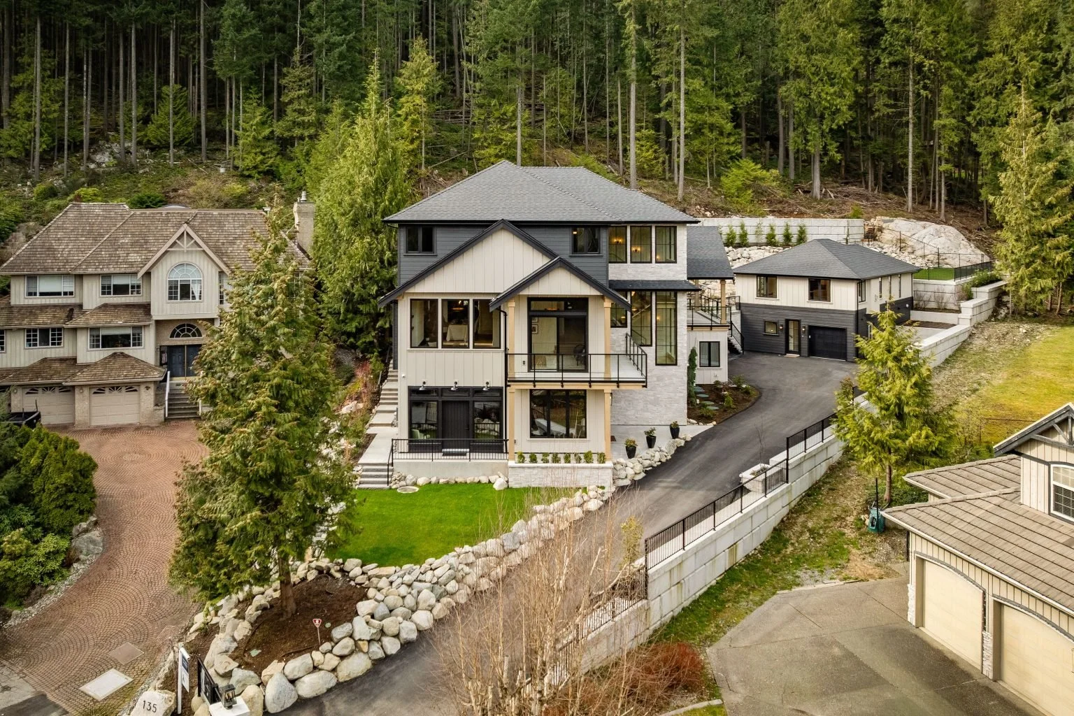 Modern multi-story house with a driveway and front yard, surrounded by trees and neighboring houses in a hilly residential area.