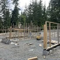Construction site with wooden framing for a building, surrounded by trees and gravel ground.