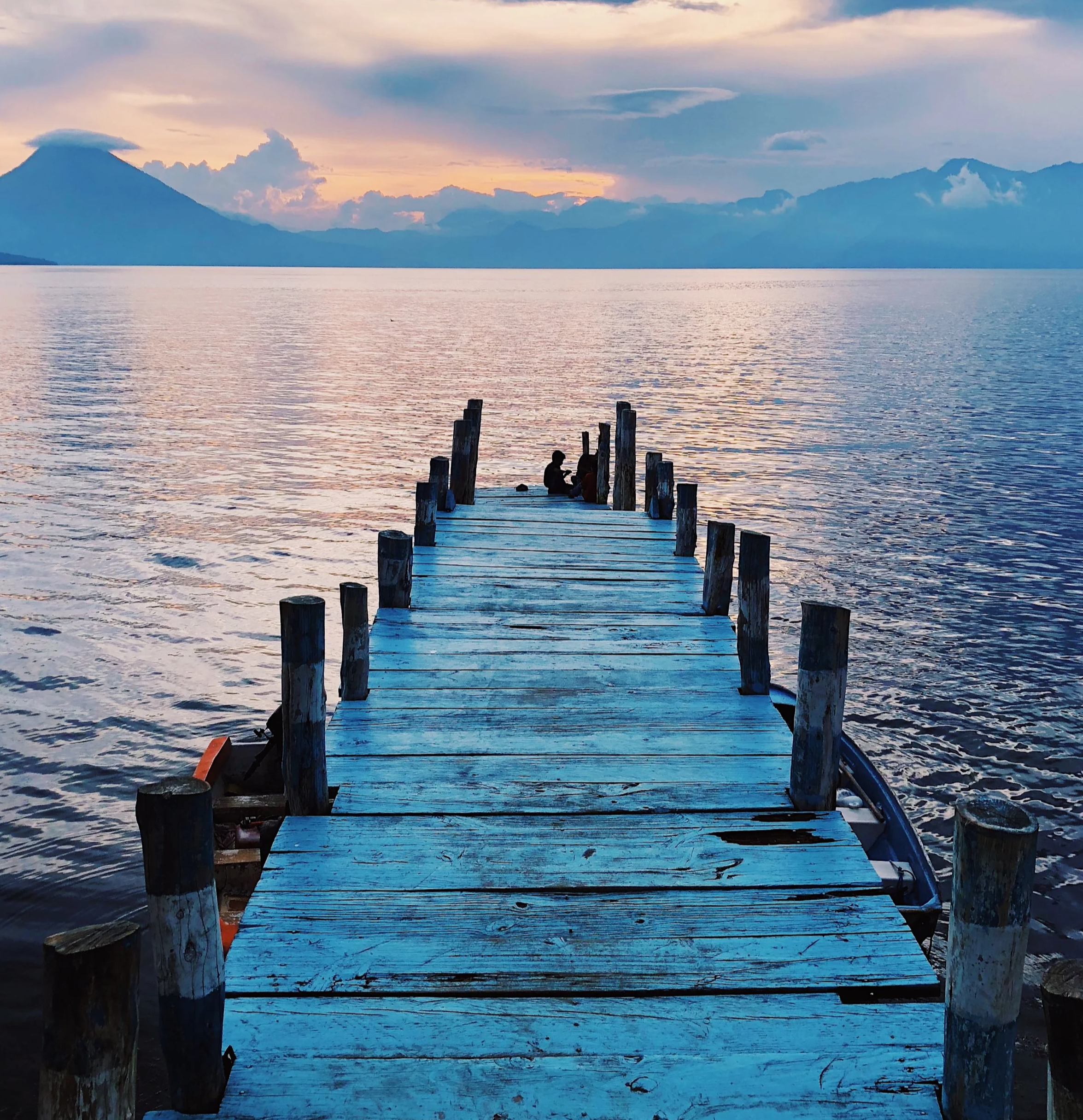 A wooden dock extending into a calm body of water with a mountain range in the background during sunset.