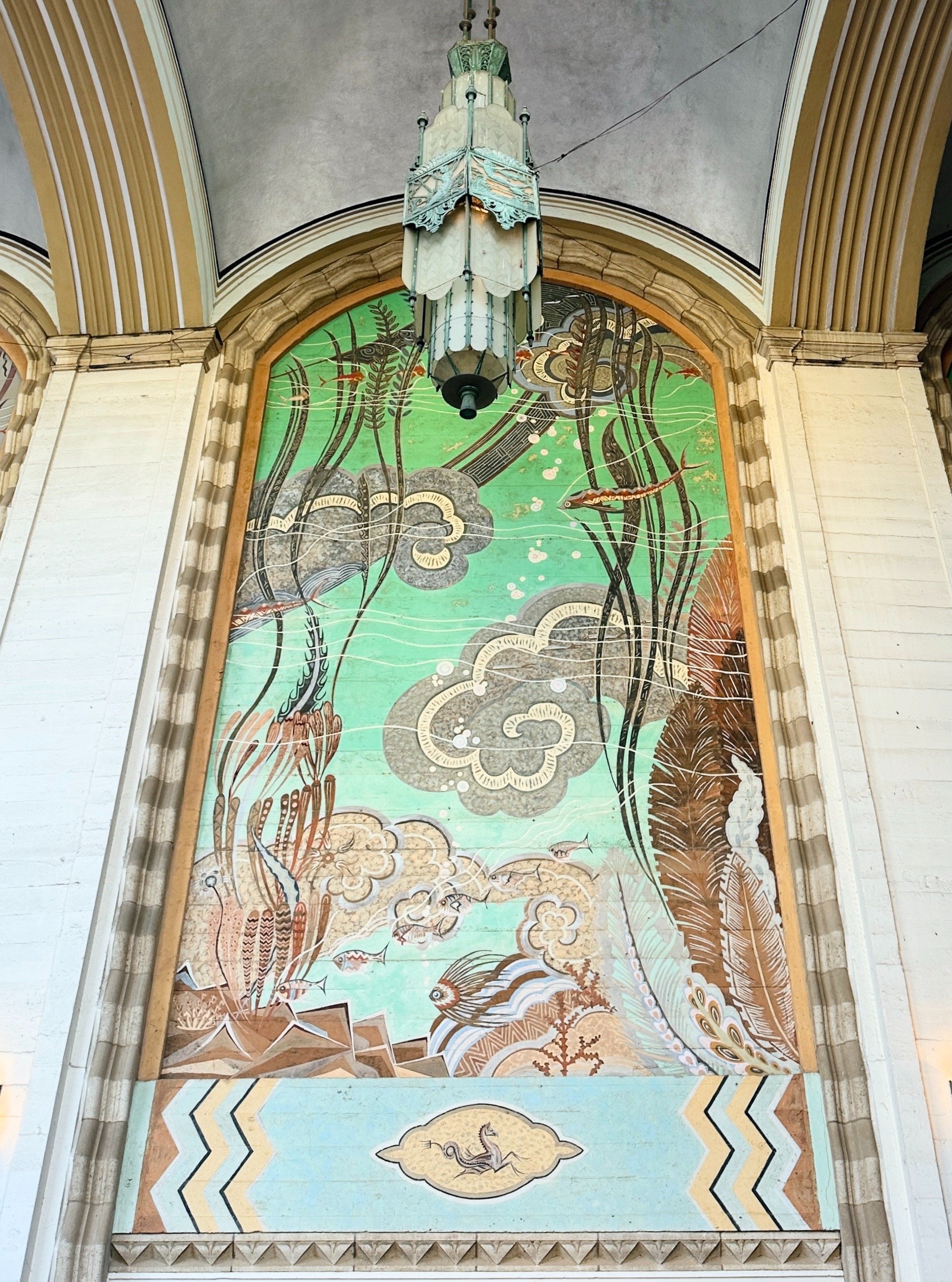 A large mural of an underwater scene with fish, seaweed, and clouds, painted on a wall inside a building with beige arches. There is a hanging lantern-style light fixture above the mural.