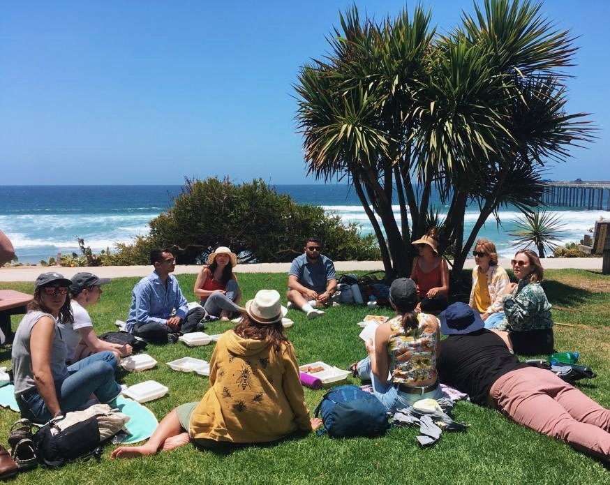 Group of people having a picnic or outdoor meeting on grass near the beach, sitting around a large tree with the ocean and a pier in the background.