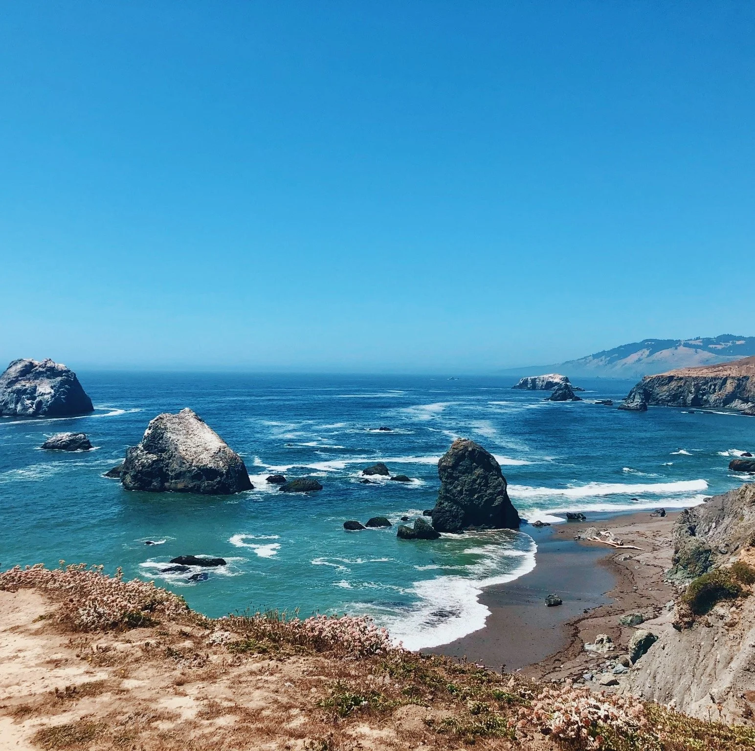 Coastal landscape with large rocks in the ocean, sandy shore, cliffs on the right, and a clear blue sky.