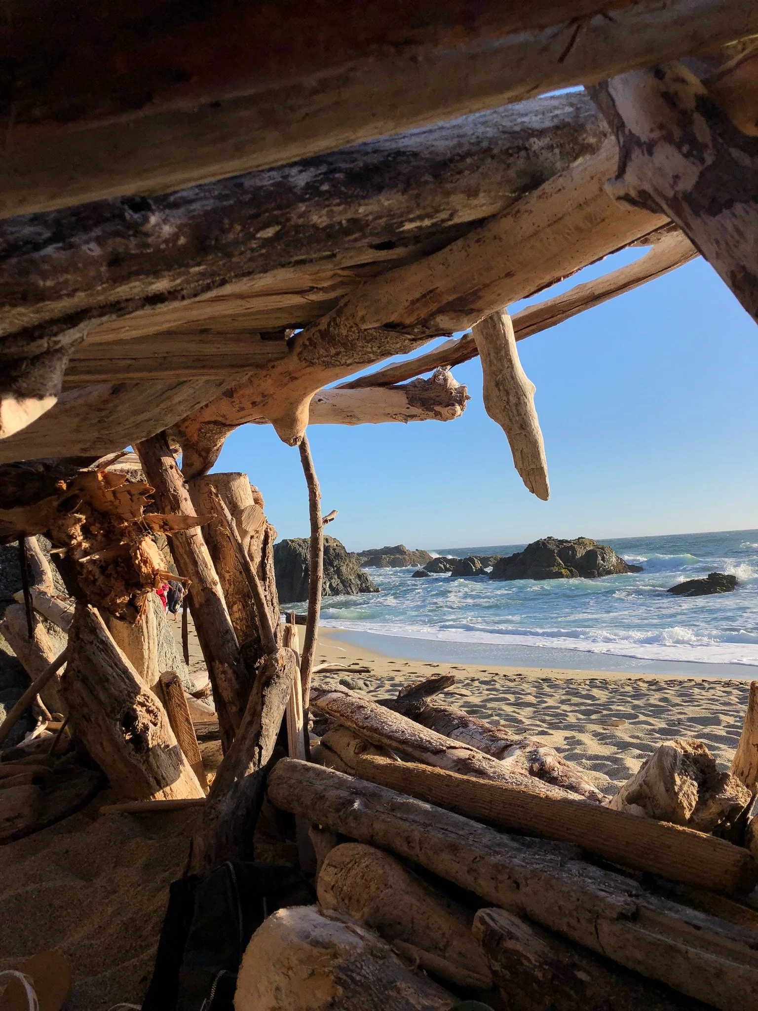 View of a beach through a driftwood shelter with rocks and ocean waves in the background