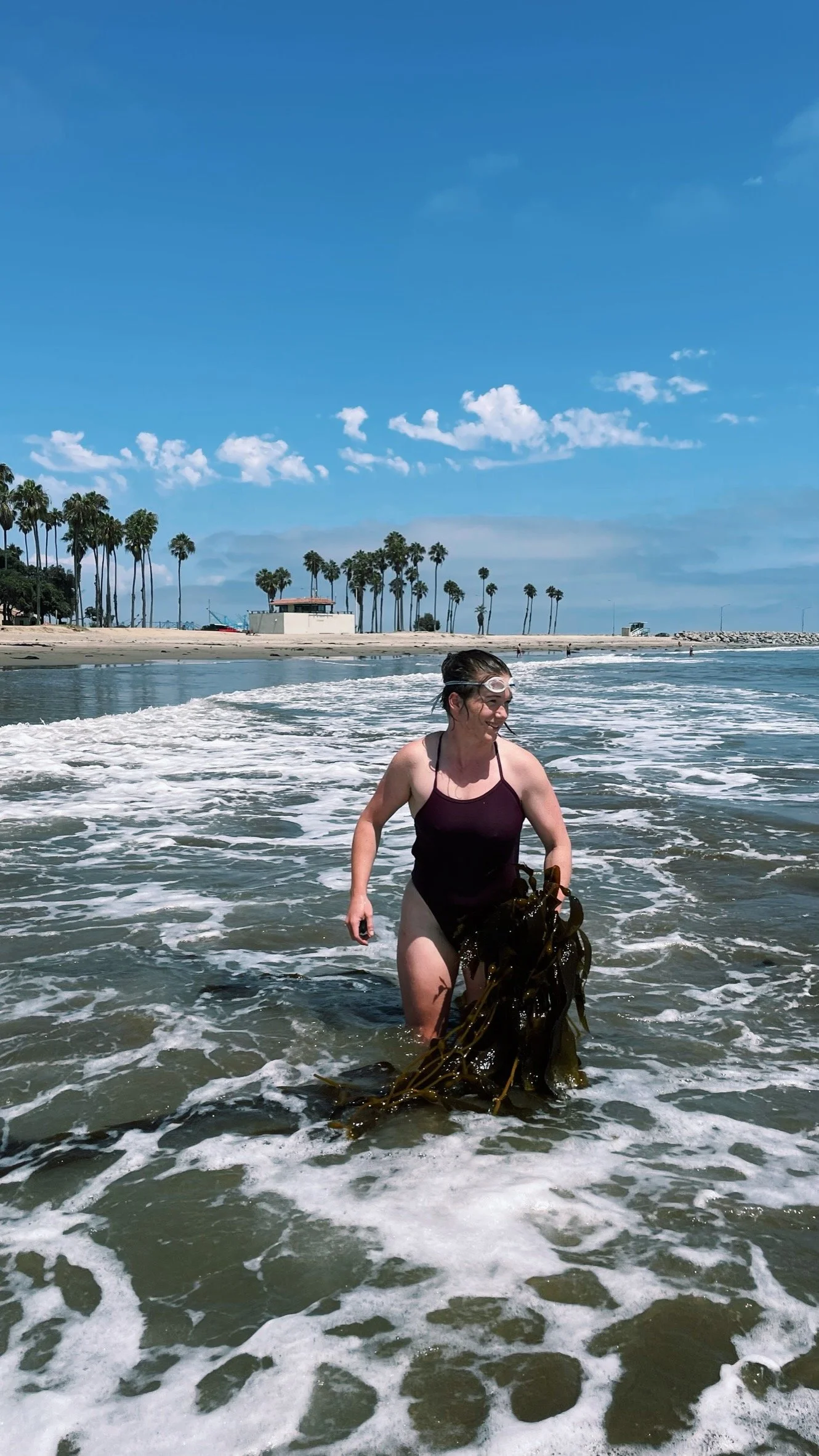Woman in a black swimsuit walking in the ocean with seaweed, wearing goggles on a sunny beach, palm trees, and a blue sky in the background.