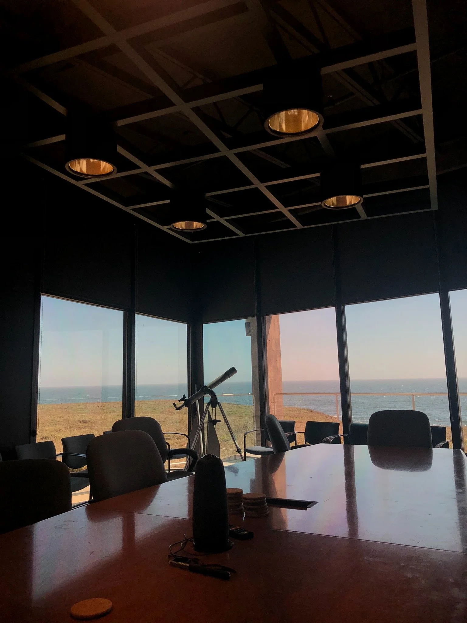 Conference room with large windows overlooking the ocean, including a telescope, chairs, and a table with various items.