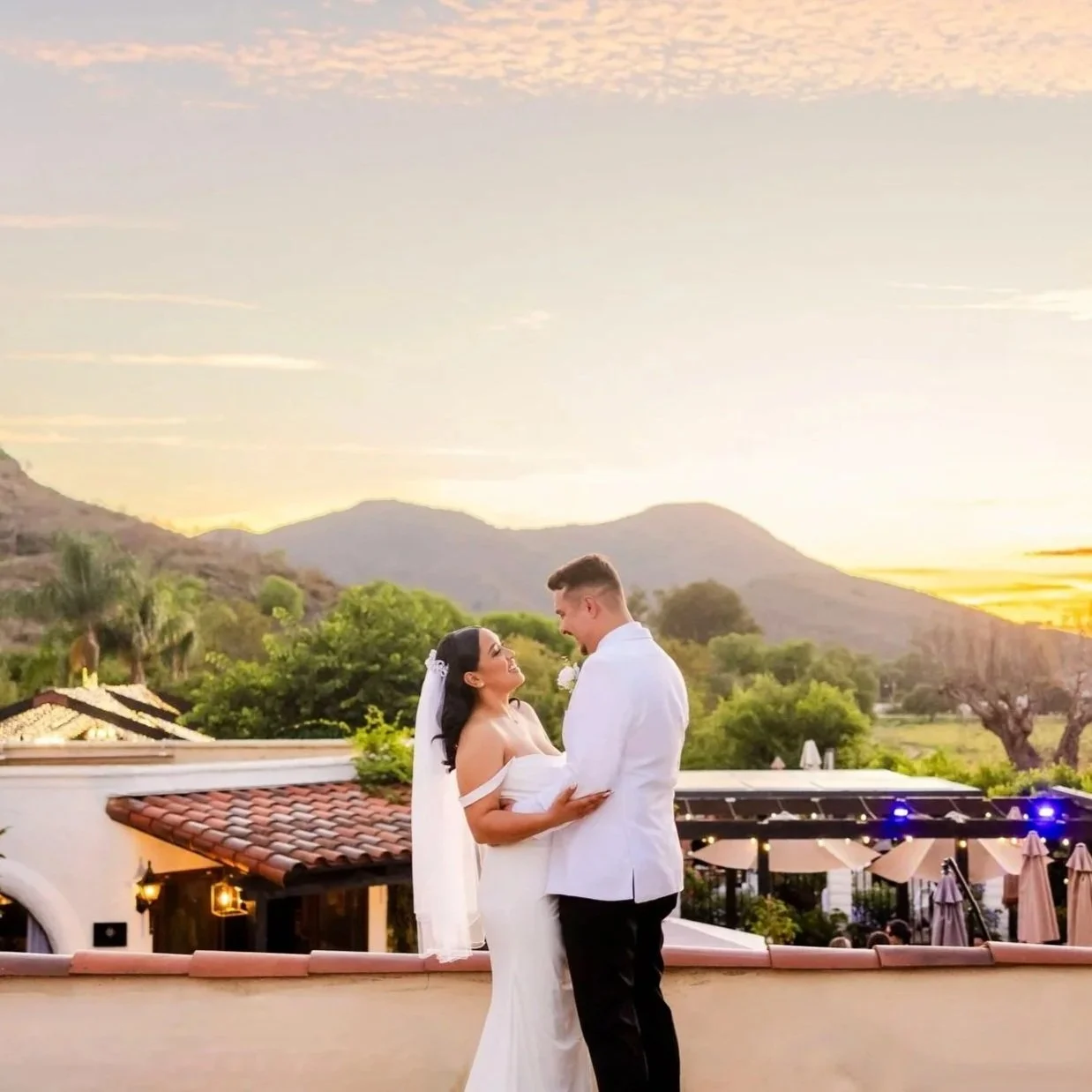 Bride and groom in wedding attire sharing a tender moment on a balcony at sunset, with mountains and trees in the background.