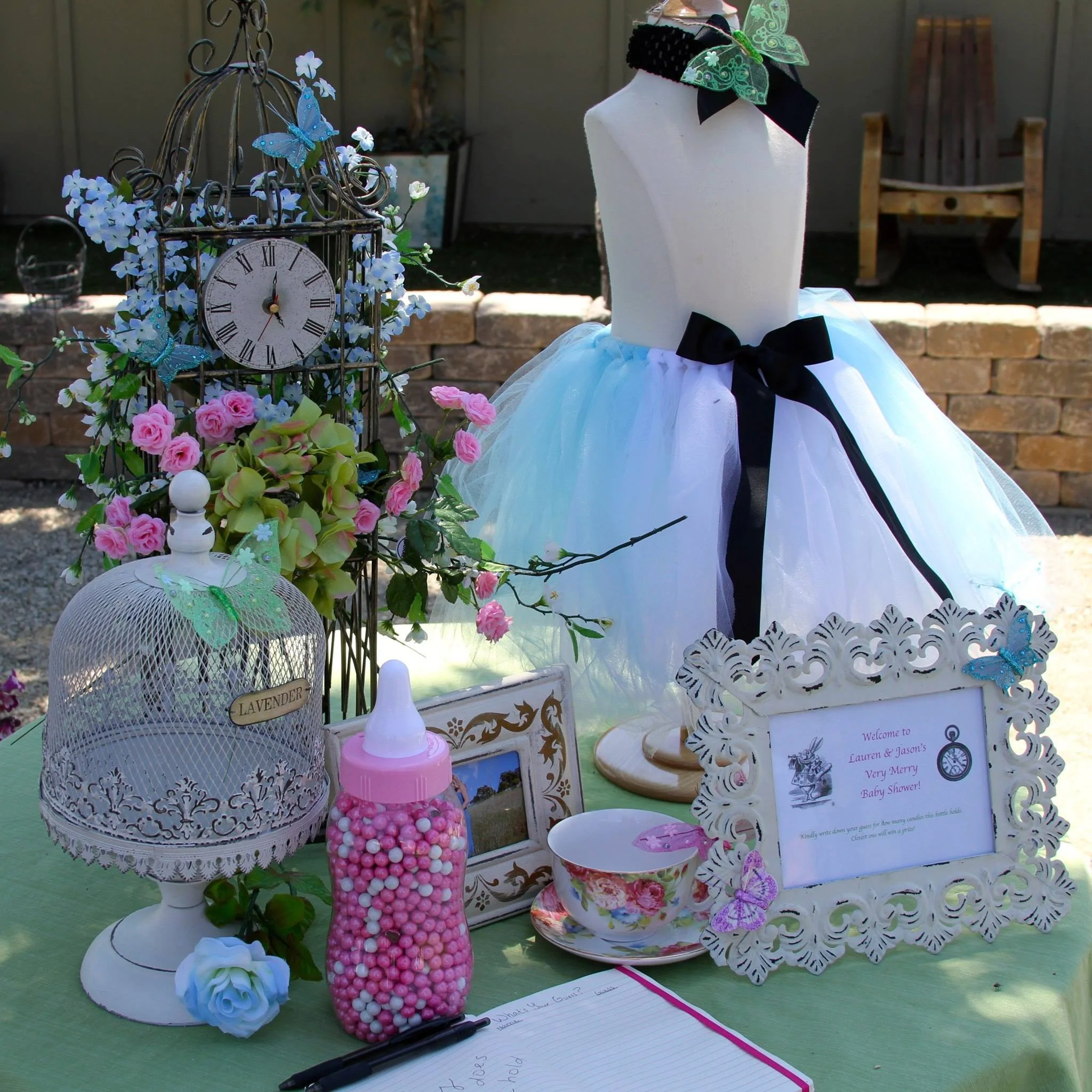 A children's baby shower table decorated with a mannequin dressed in a white tutu with black ribbon, framed sign, teacup and saucer, vintage style birdcage, pink and white candy bottle, artificial flowers, and decorative items including a clock, butt