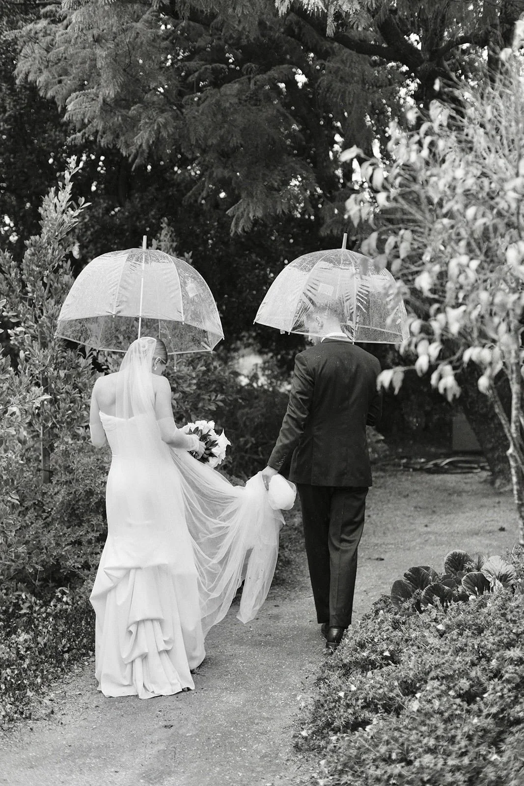 Black and white photo of a bride and groom walking together outdoors with umbrellas, the bride in a wedding gown holding flowers, and they are surrounded by trees and bushes.