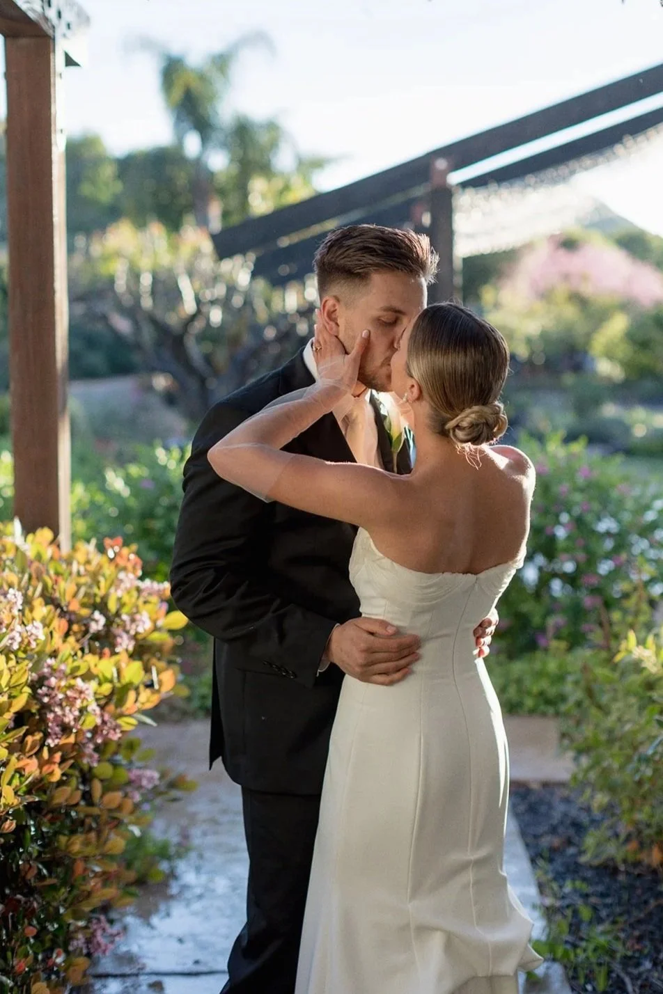 A bride and groom kissing outdoors on their wedding day, surrounded by greenery and flowers.