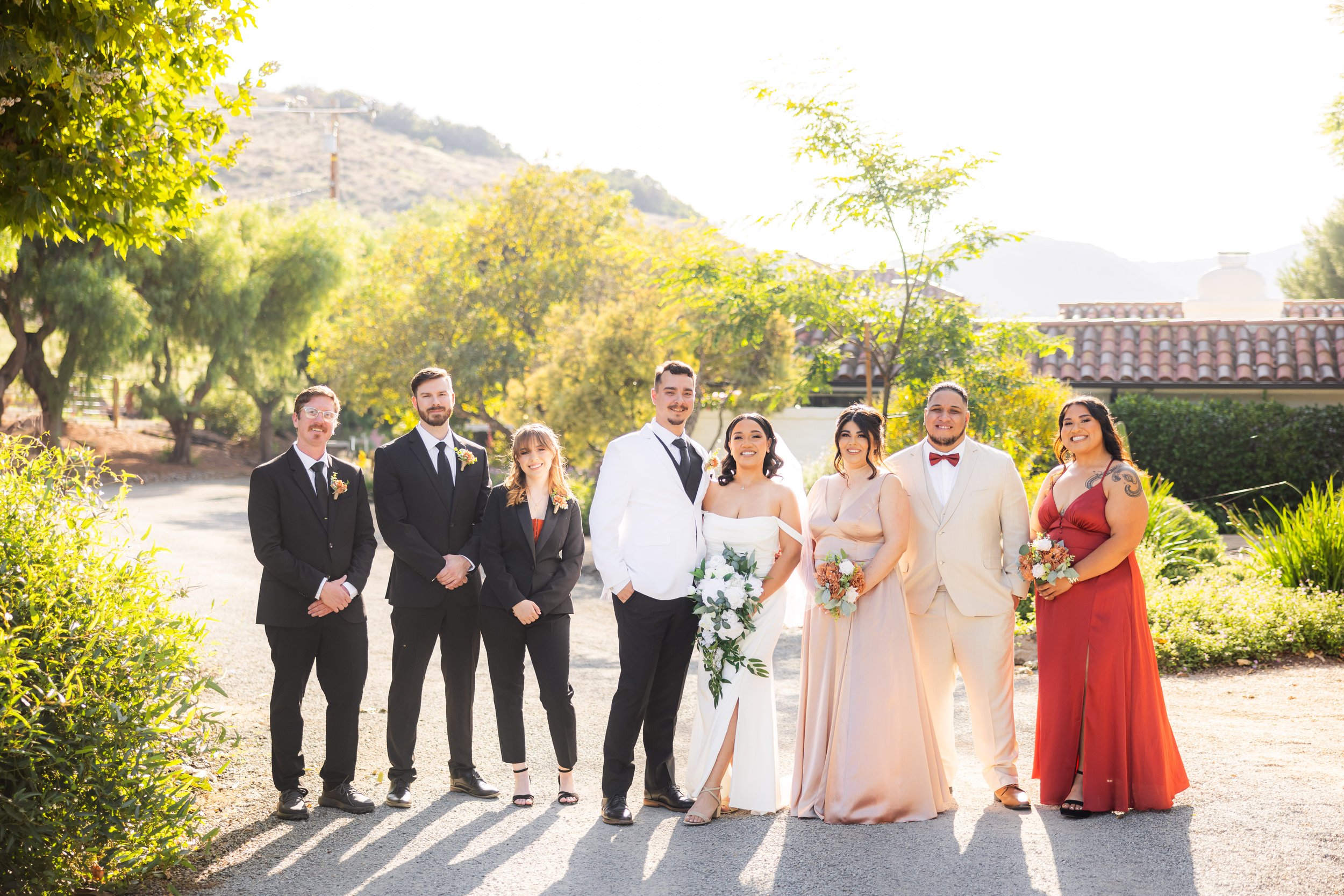 Group of eight people dressed in formal attire standing outdoors on a sunny day, with trees and a house with tile roof in the background, celebrating a wedding.