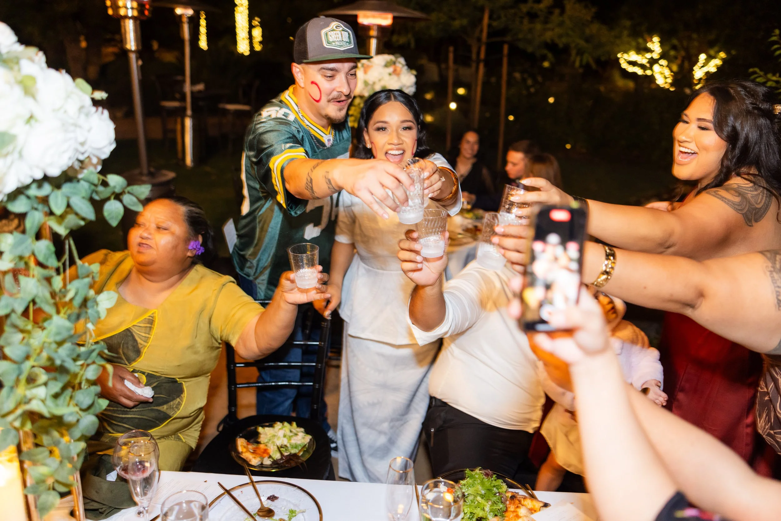 People celebrating at a party, raising glasses for a toast, with a woman in a white dress in the center, surrounded by friends and family, with food on the table and string lights in the background.