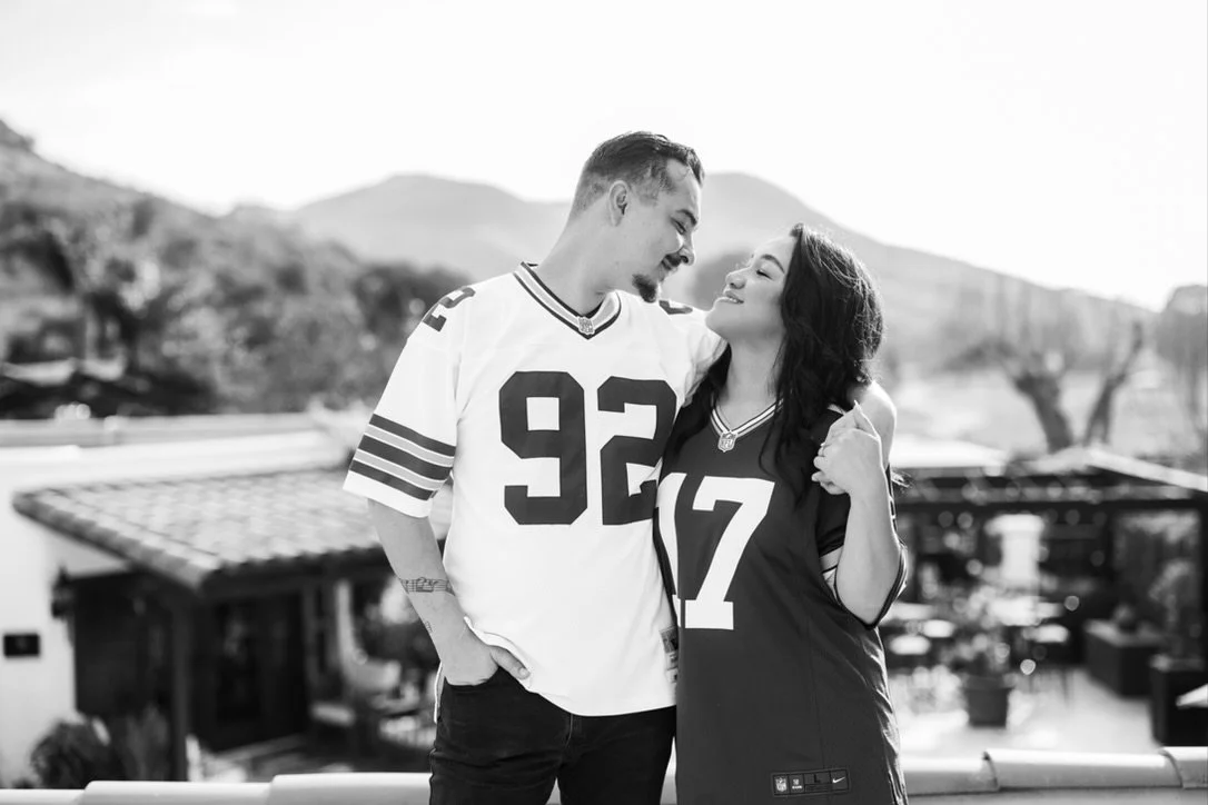 A young couple stands close together outdoors, smiling at each other, with a scenic background of mountains and trees with jerseys on.