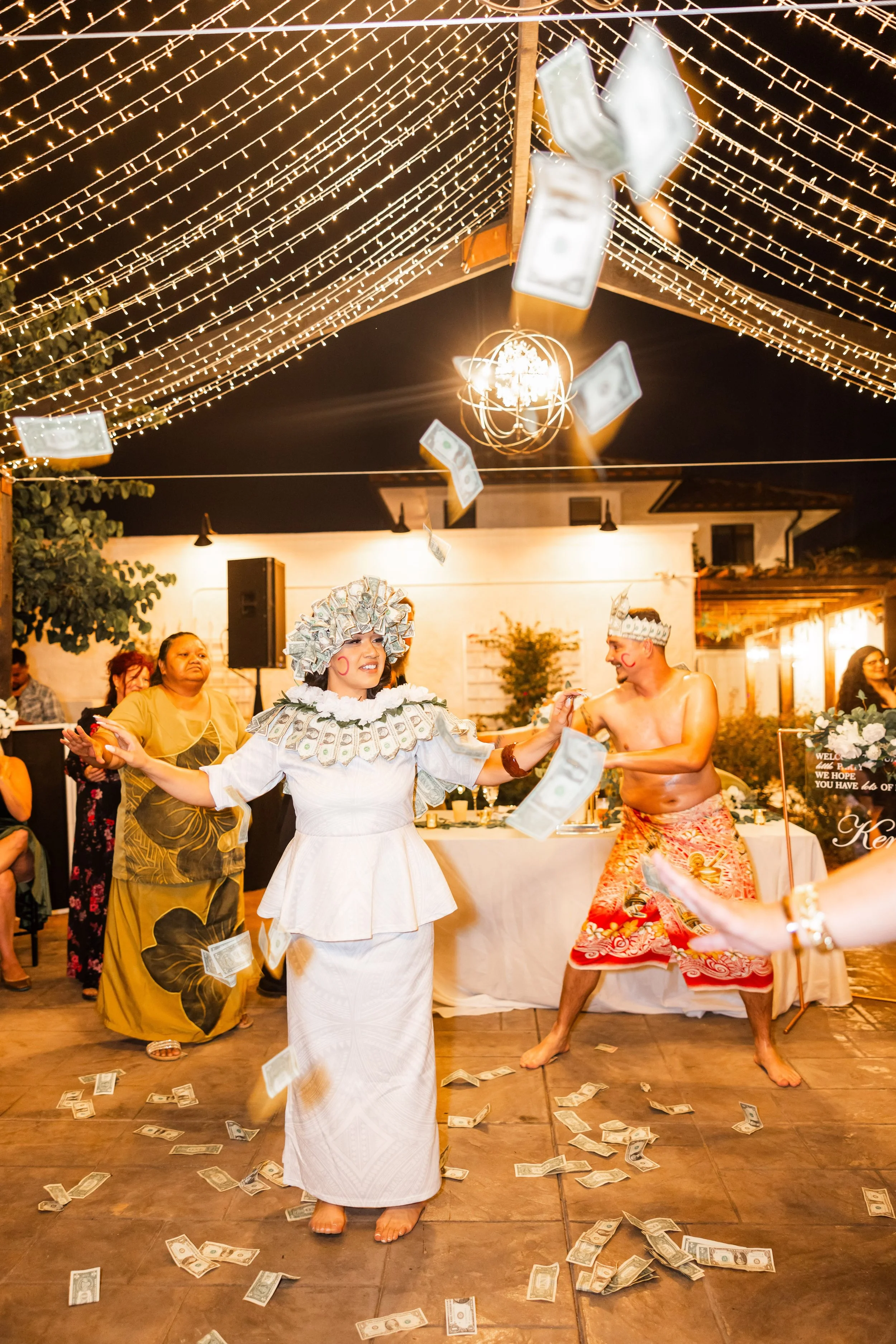 People celebrating at a party under string lights with money falling from above. A woman in a white dress and elaborate headpiece stands in the center, surrounded by others, some in traditional costumes, with a lively atmosphere.
