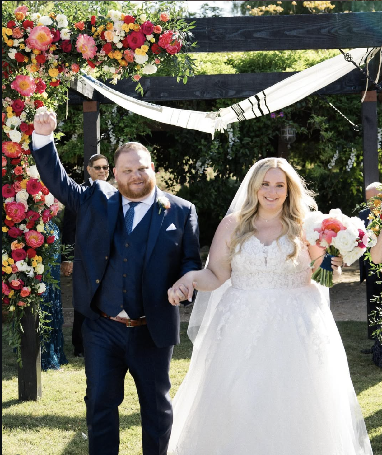 A newlywed couple walking hand in hand after their wedding ceremony outdoors, with a floral arch and decorations in the background, celebrating their marriage.