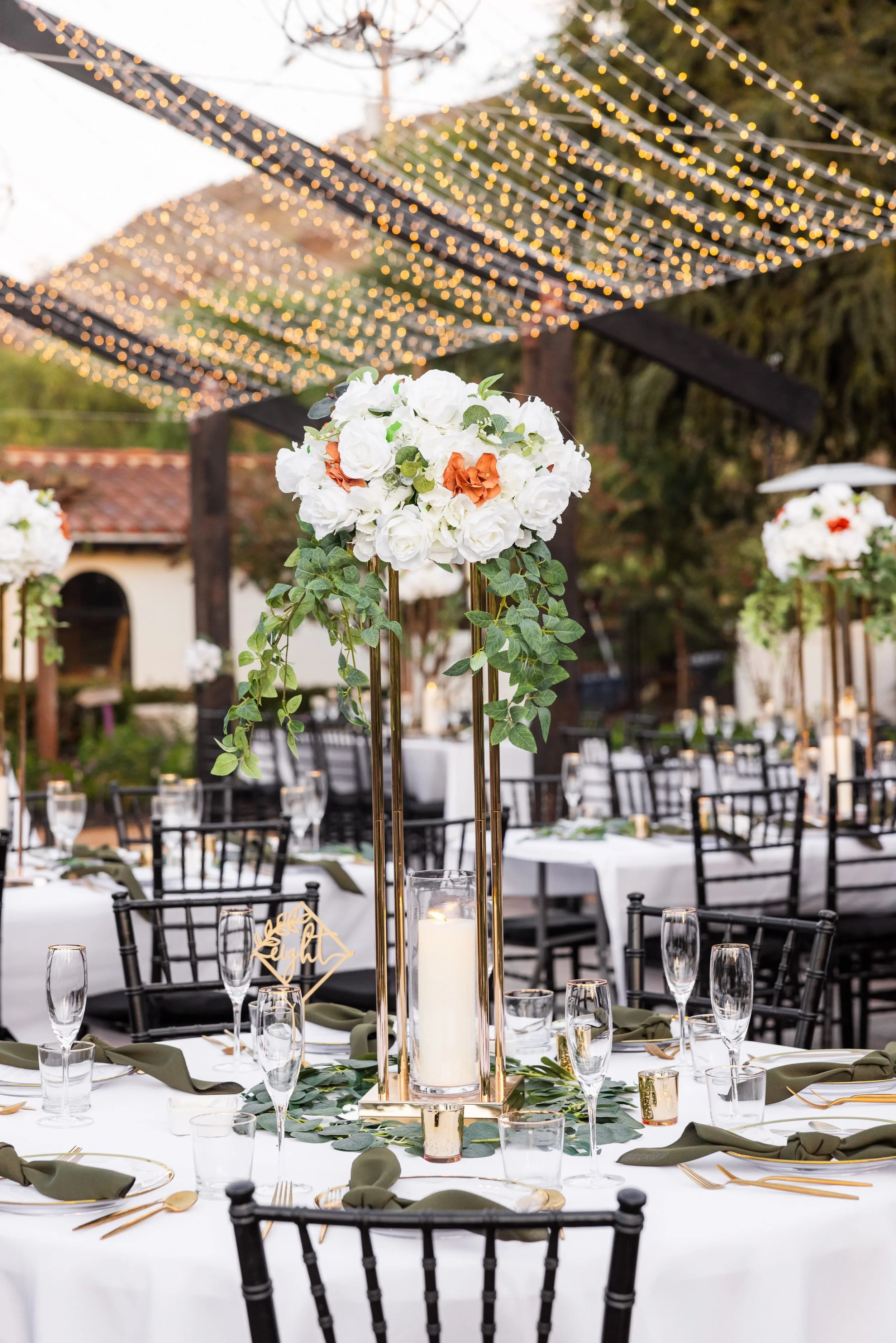A decorated outdoor event table with floral centerpiece, candles, glassware, and green napkins, under string lights.