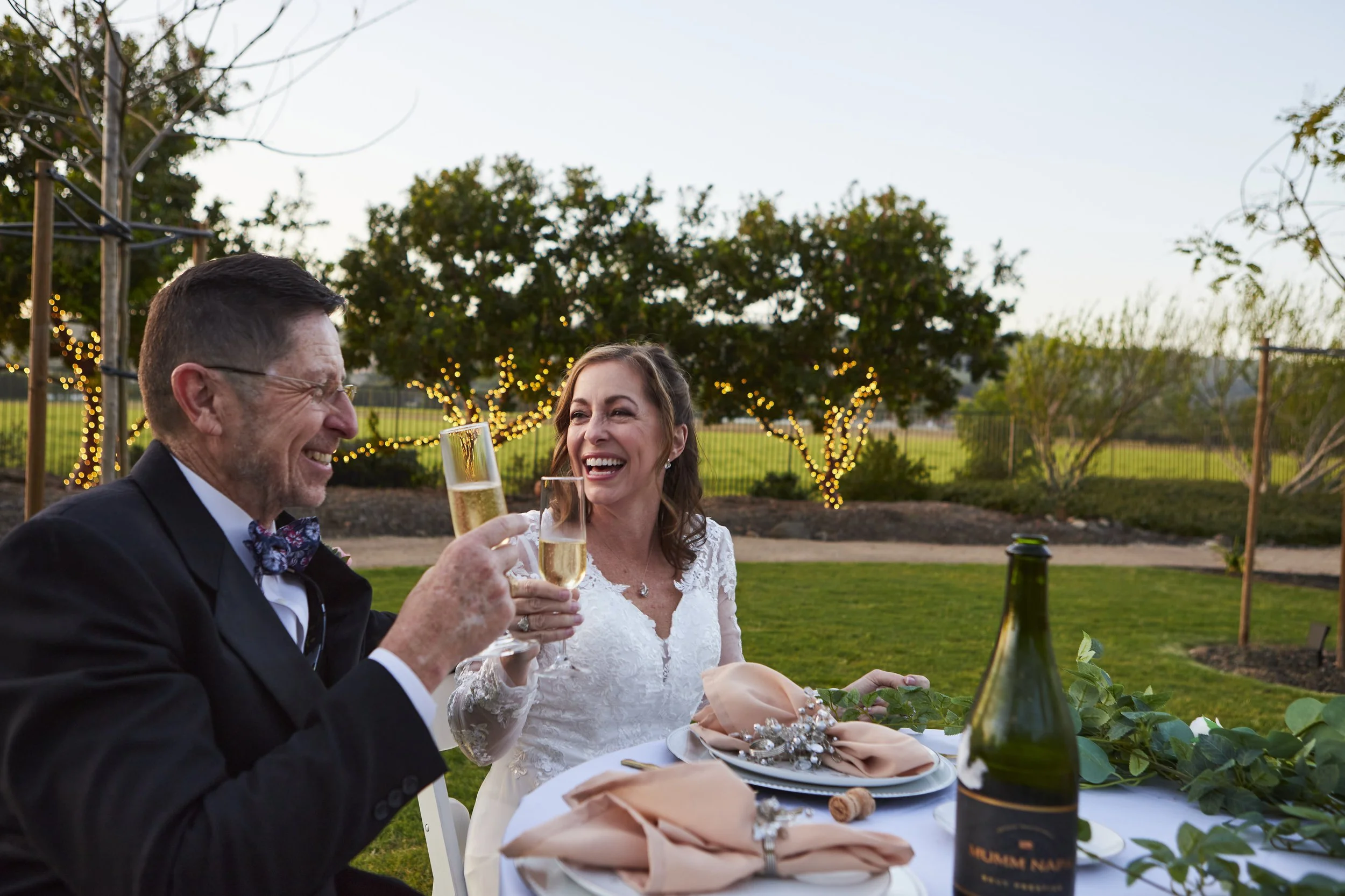 A joyful wedding reception outdoors with a bride and groom toasting with champagne glasses during sunset. The groom is wearing a black tuxedo with a bow tie, and the bride is in a lace wedding dress. A decorated wedding cake and wine bottle are on th