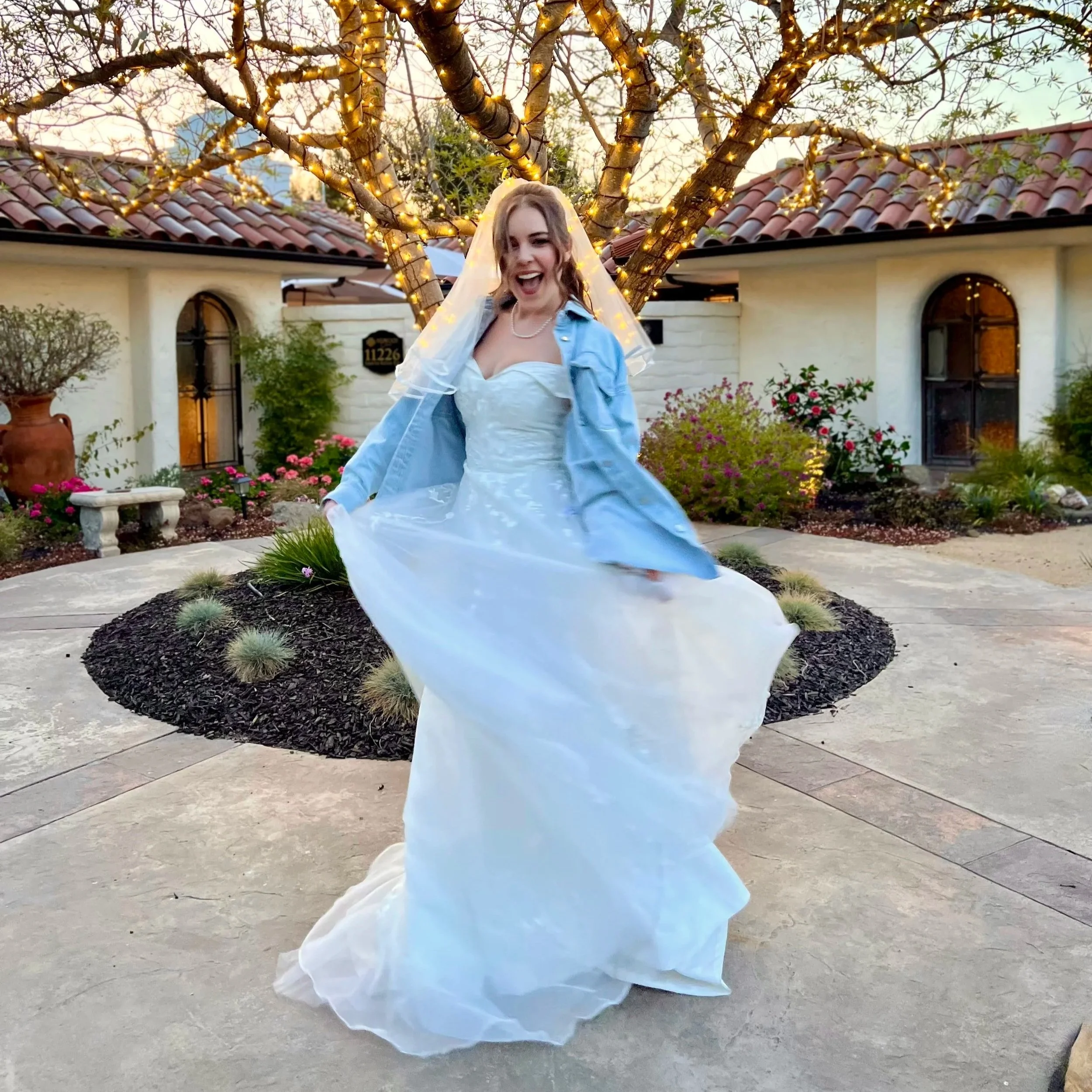 A woman in a wedding dress and blue jean jacket cheerfully dancing outdoors in front of a decorated tree with string lights, with a house in the background.