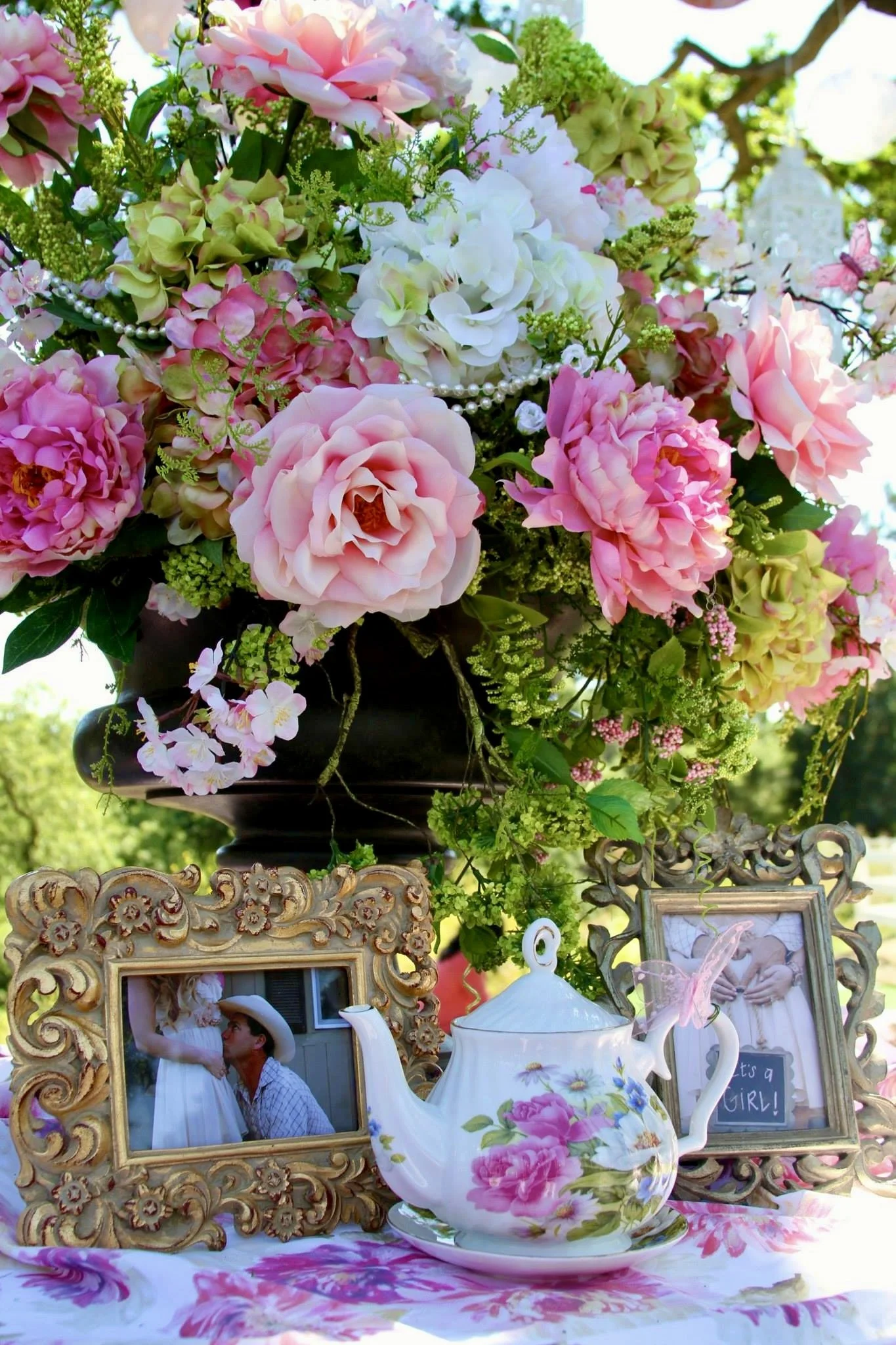 A floral arrangement of pink, white, and green flowers in a black vase, with picture frames, a floral teapot, and decorative items arranged on a table with a pink floral tablecloth.