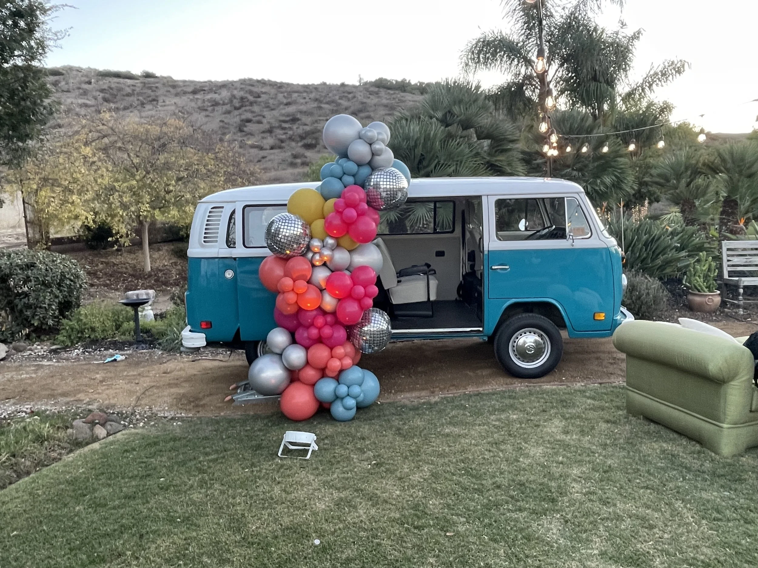 A vintage blue and white van decorated with a large cluster of colorful balloons, including pink, red, blue, silver, and gold, standing outdoors in a park with trees, grass, and string lights hanging above.