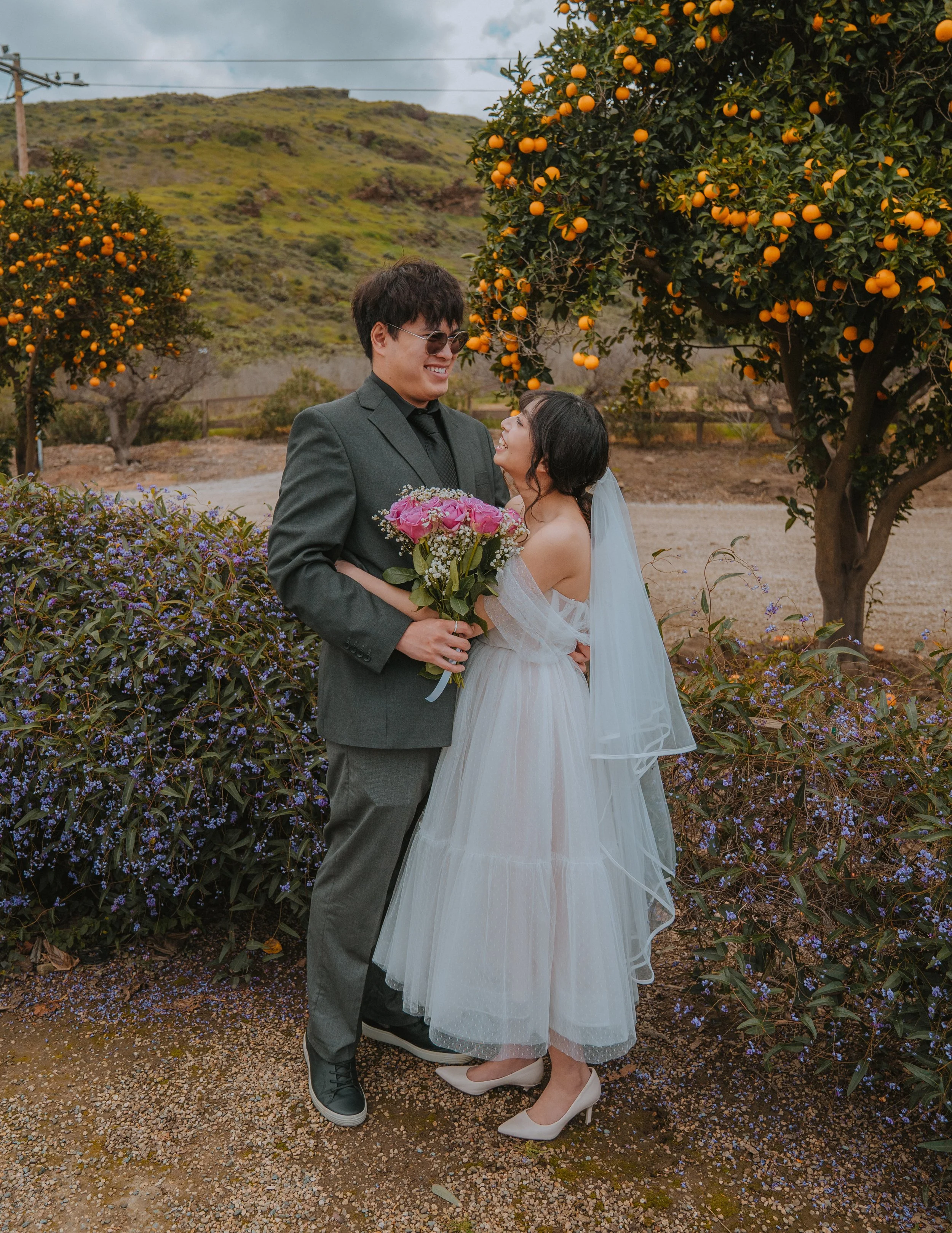 A couple dressed in wedding attire standing outdoors among orange trees and purple flowering bushes, holding flowers and smiling at each other.
