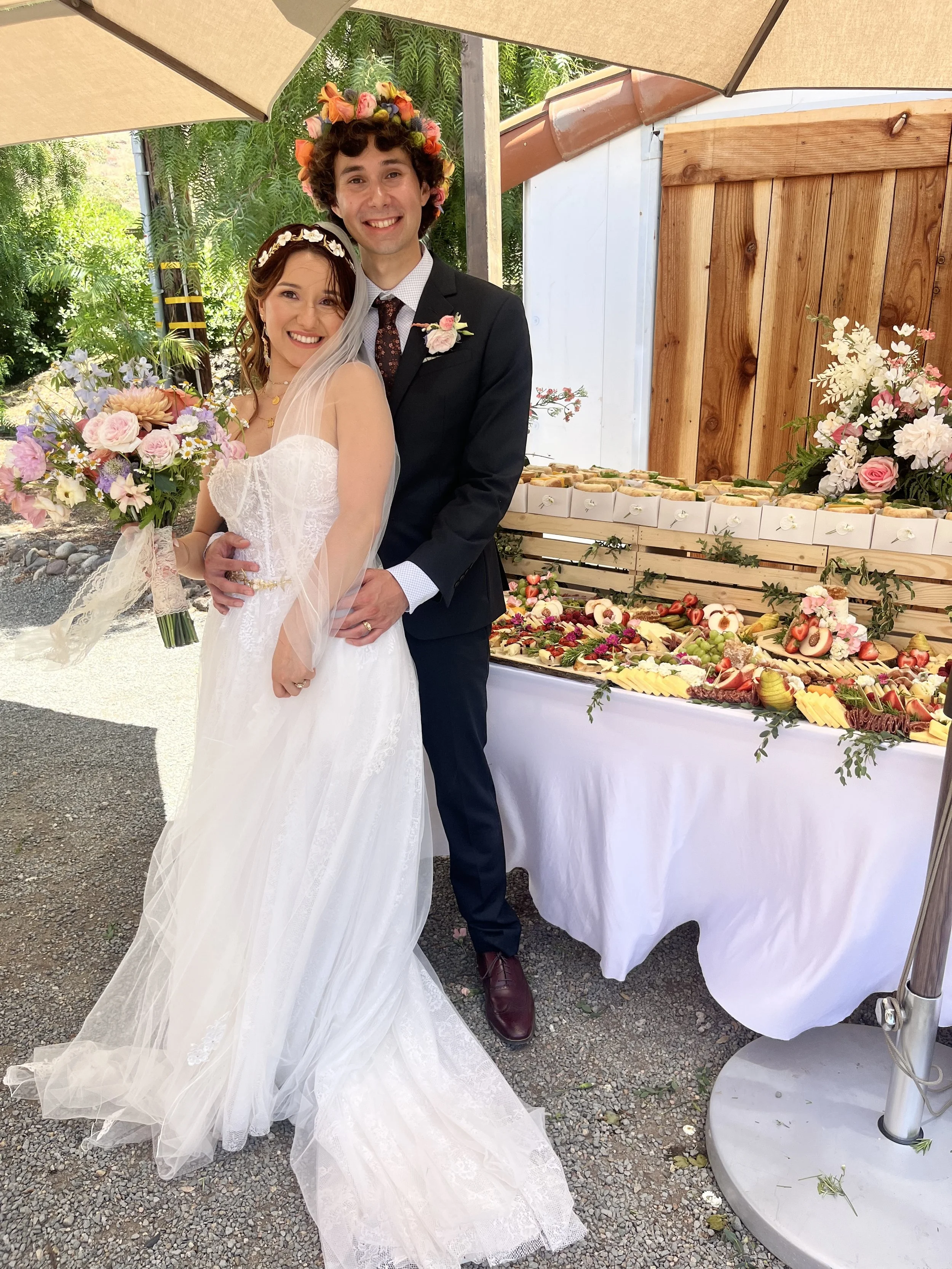 A happy bride and groom standing outside under a large umbrella, with a wedding reception table behind them filled with food and floral arrangements. The bride is holding a bouquet of flowers, and both are smiling at the camera.