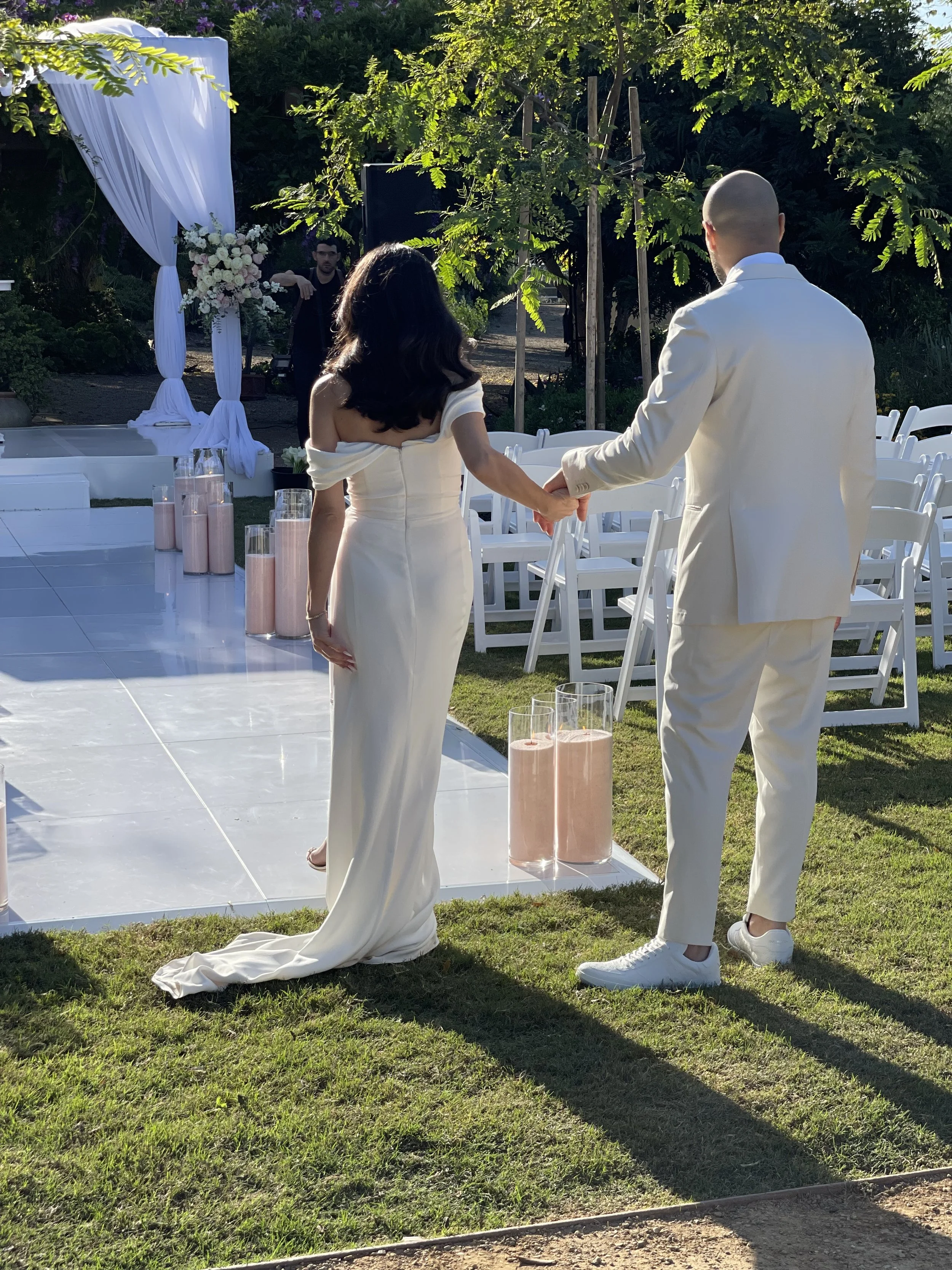 A couple holding hands during their wedding ceremony outdoors, with the woman in a white gown and the man in a white suit, standing on a grassy area with chairs and flowers in the background.